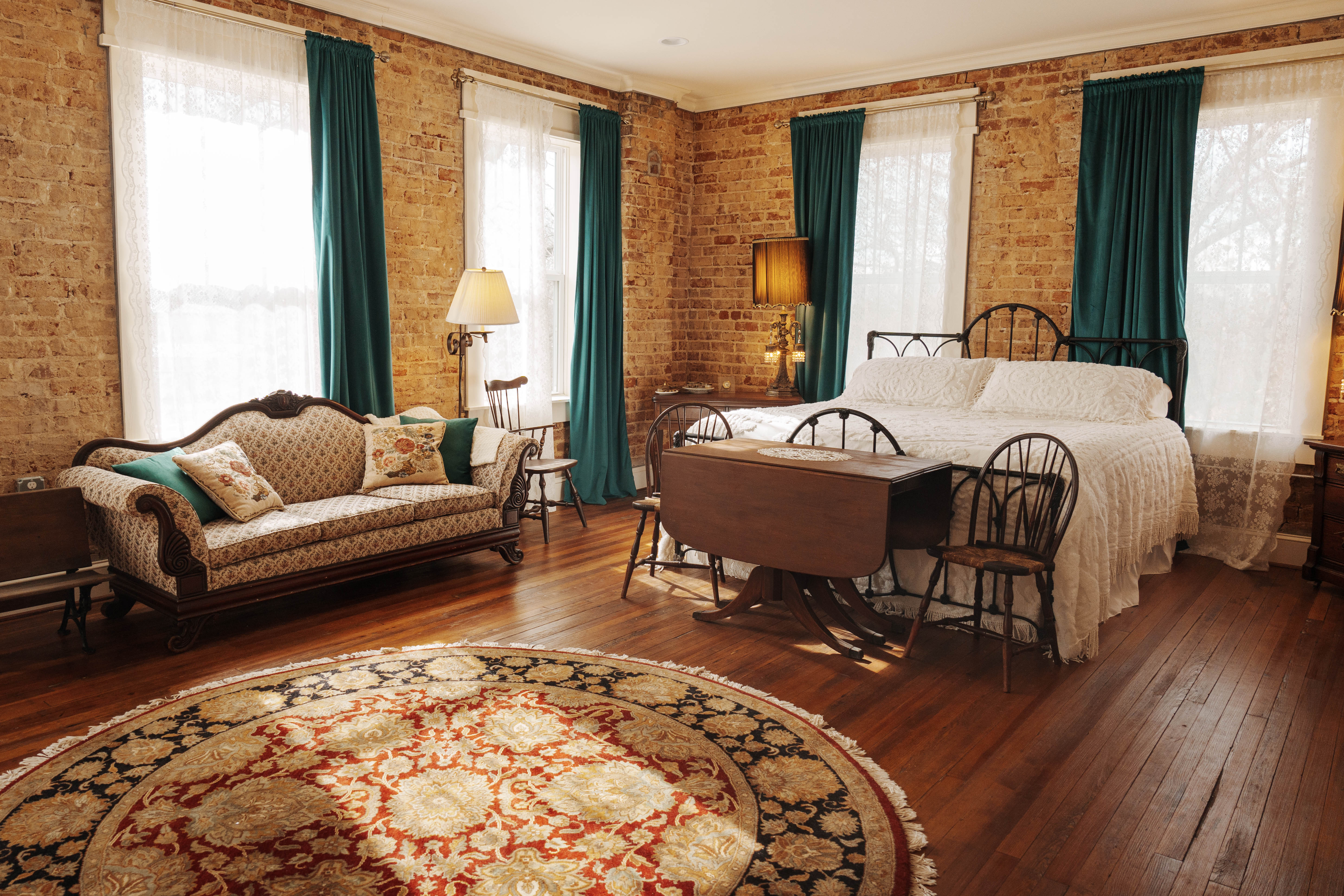 A bedroom with a king-size bed, a beige sofa with decorative pillows, a round patterned rug, and multiple windows with teal curtains.