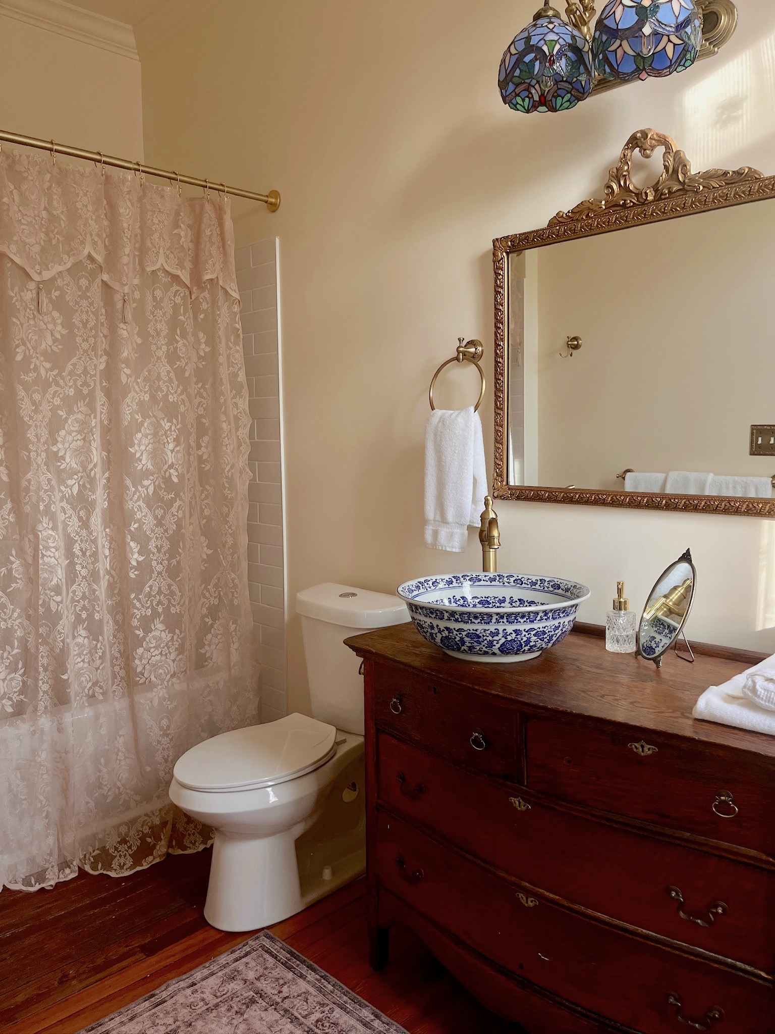 A bathroom with a dark wooden dresser-style vanity, a blue and white patterned bowl sink, a white toilet, and a shower with a lace curtain.