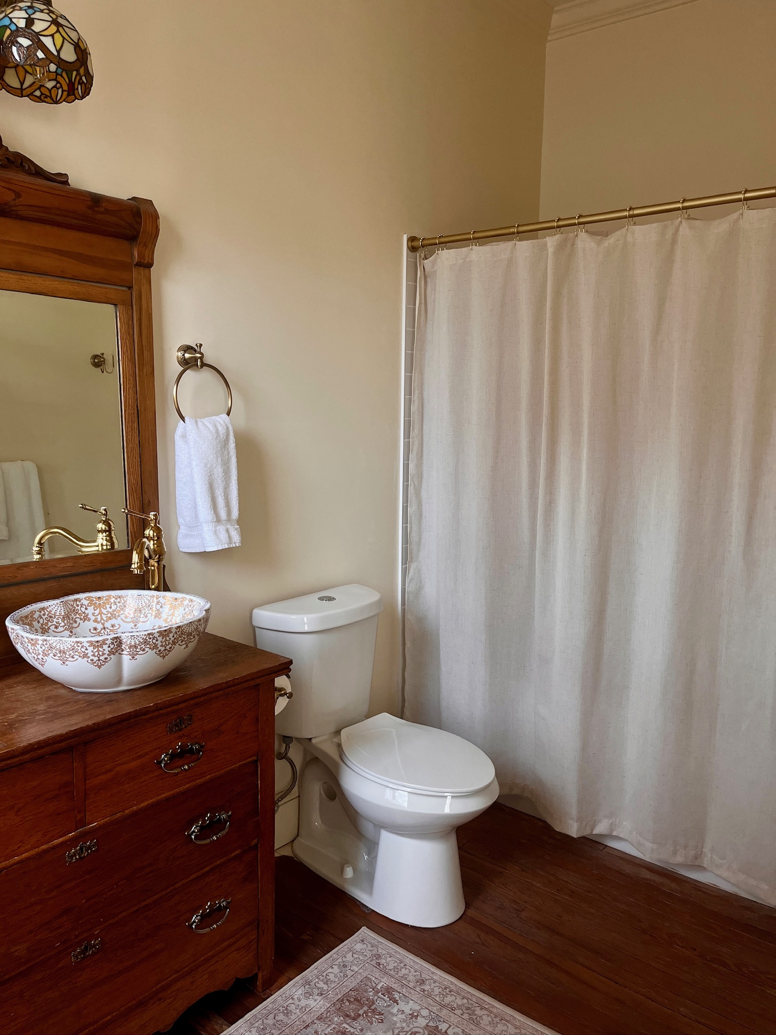 Bathroom with dresser-style vanity with floral patterned bowl sink, wooden framed mirror, toilet, and shower.
