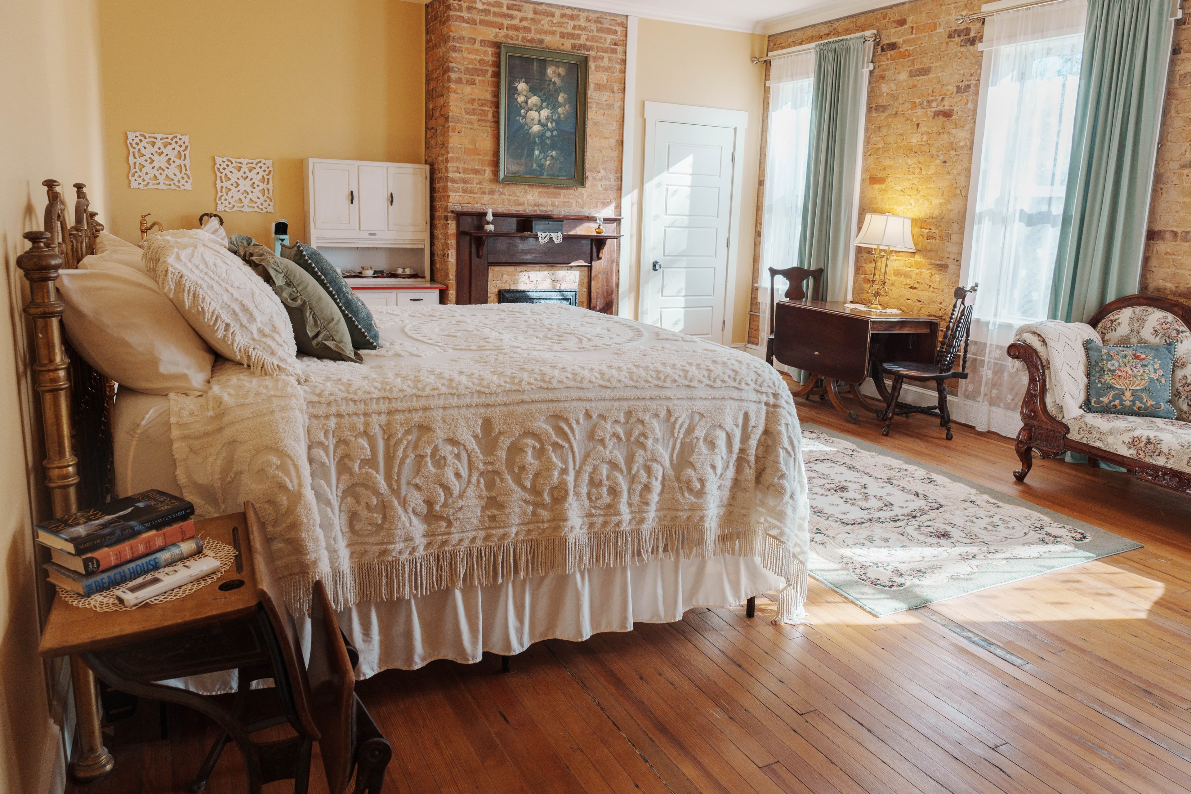 A romantic bedroom featuring a king-size bed with dreamy bedding, a wooden drop-leaf table with two chairs, a small sofa with patterned pillows, and a brick fireplace. The room has large windows with sheer curtains and a wooden floor.