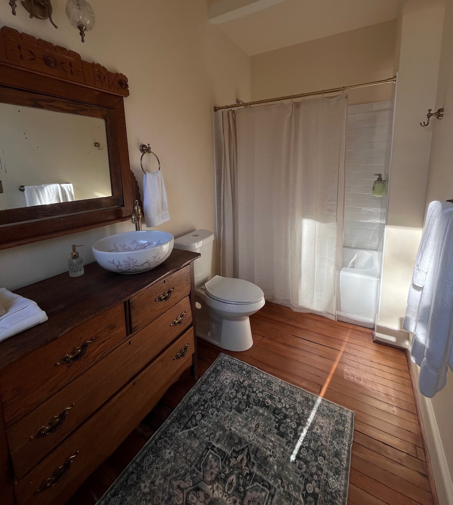 A bathroom with a wooden dresser-style vanity, a white vessel sink, a white toilet, and a shower with a white curtain, featuring a grey patterned rug.