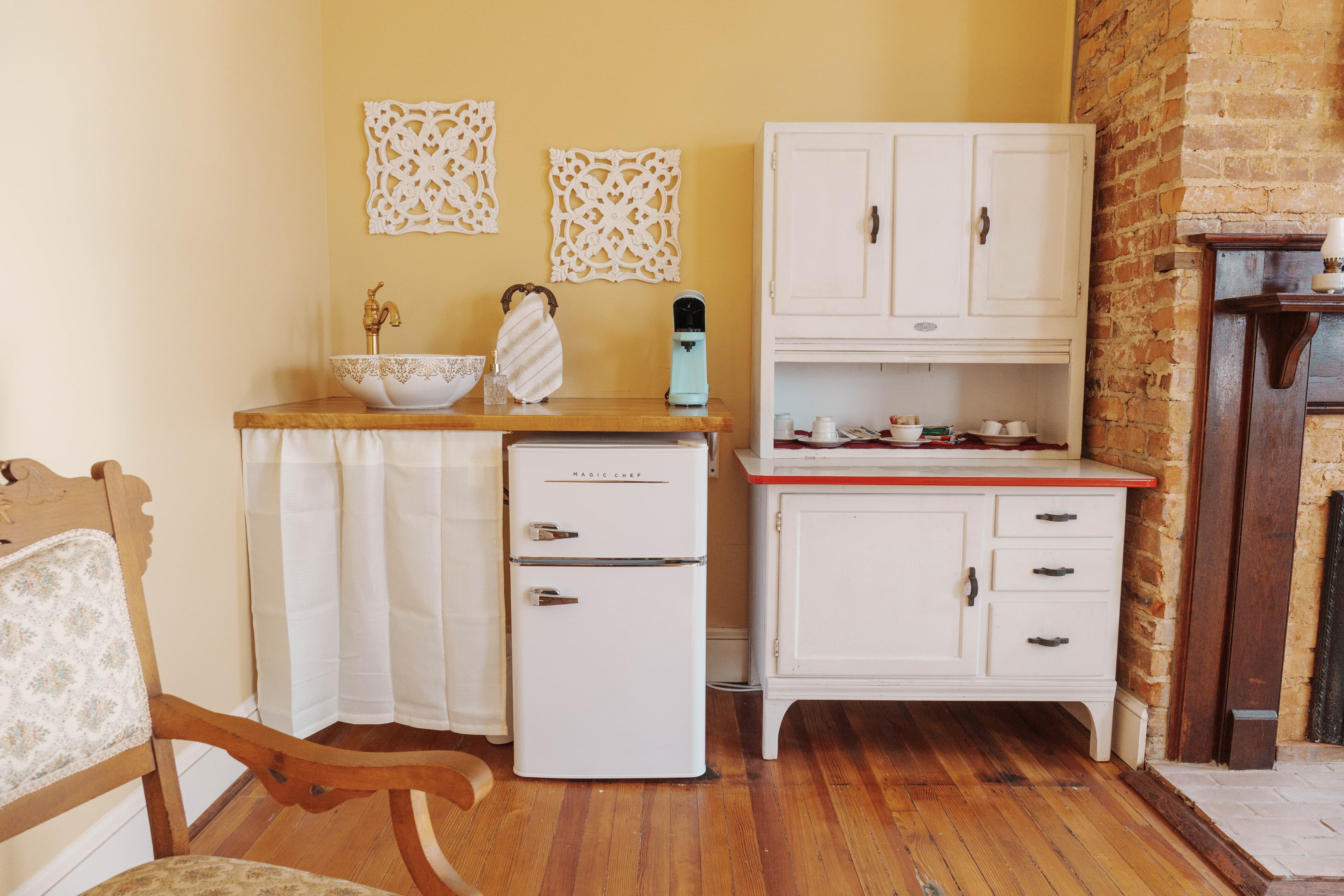 A kitchenette area with white cabinets, a mini fridge, a wooden countertop with a sink, and decorative wall hangings.