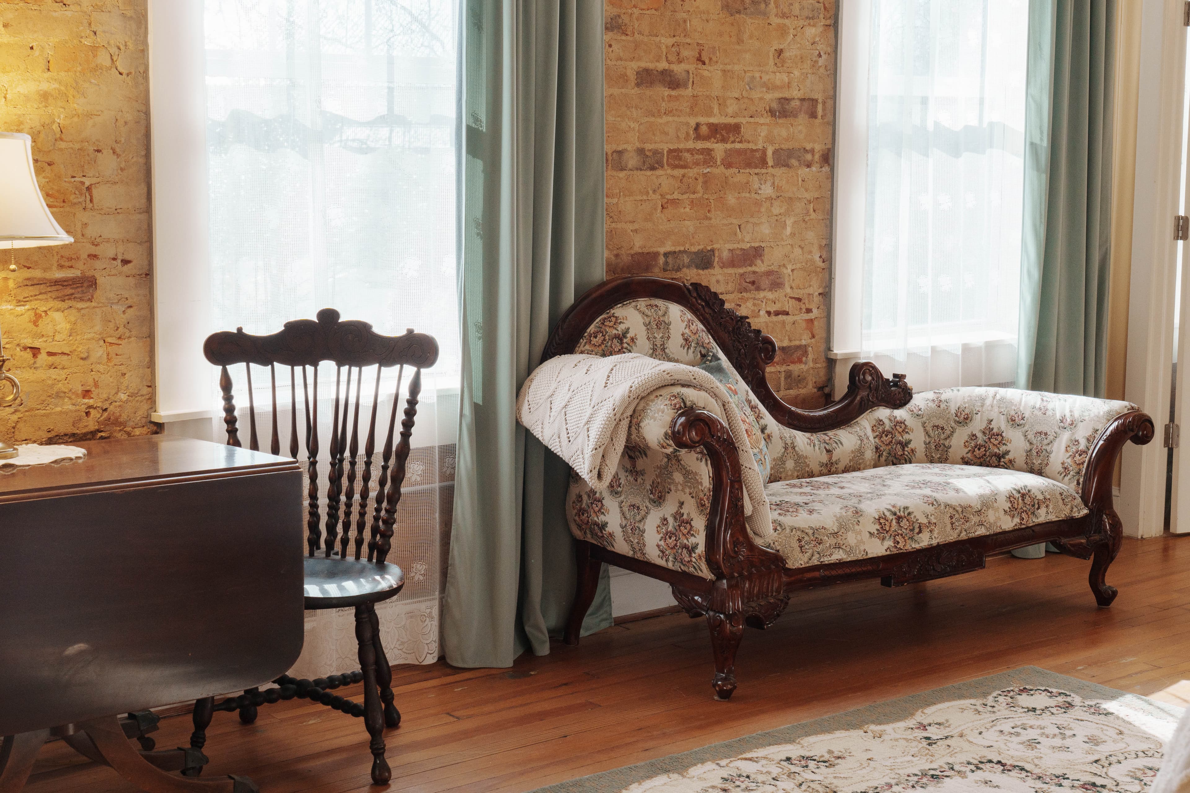 A sitting area with a floral patterned chaise lounge and a dark wooden chair near windows with sheer curtains.