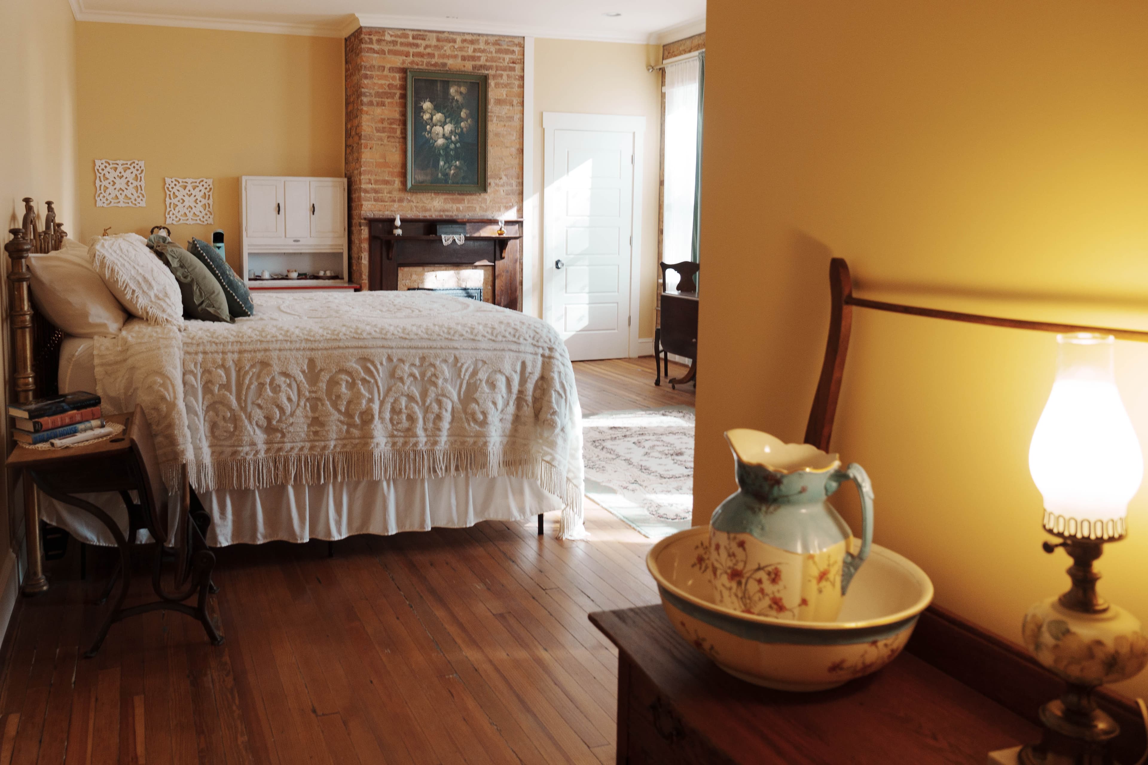 A bedroom with a bed featuring a textured white quilt, a decorative pitcher and bowl on a table in the foreground, and a fireplace with a framed picture in the background.