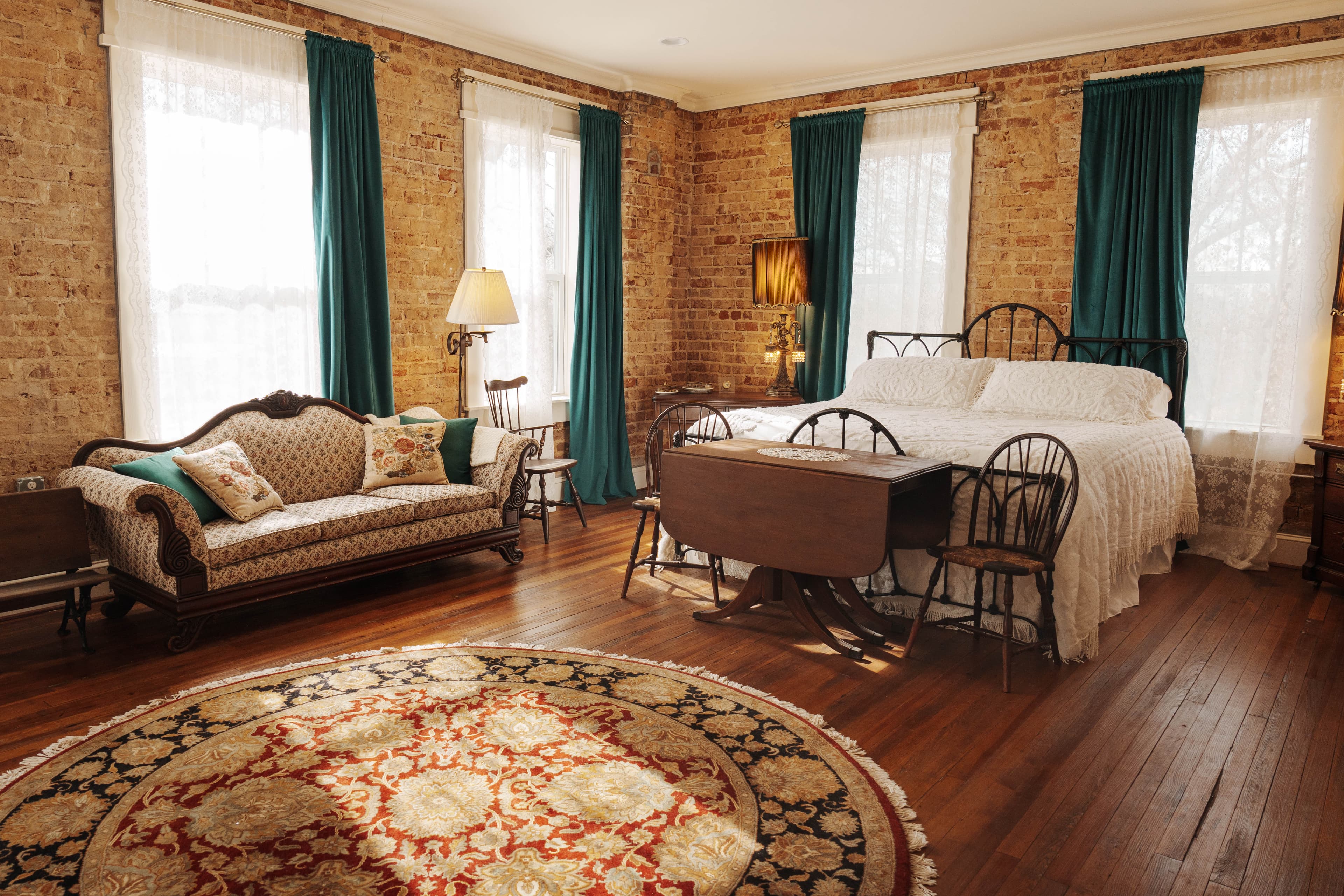 A bedroom with a king-size bed, a beige sofa with decorative pillows, a round patterned rug, and multiple windows with teal curtains.