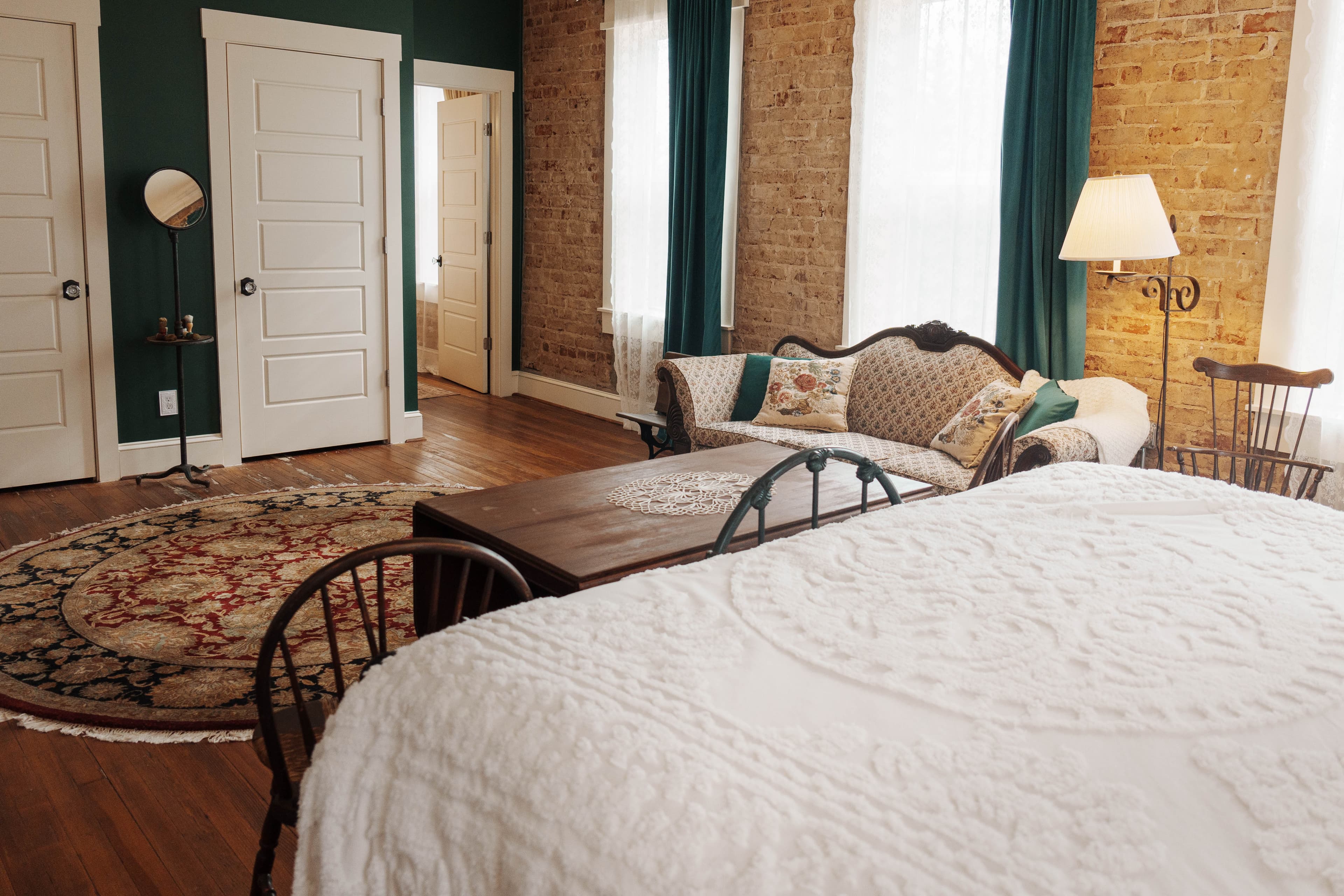 A bedroom with a white textured bed, a beige sofa with pillows, a round patterned rug, brick walls, and teal curtains.