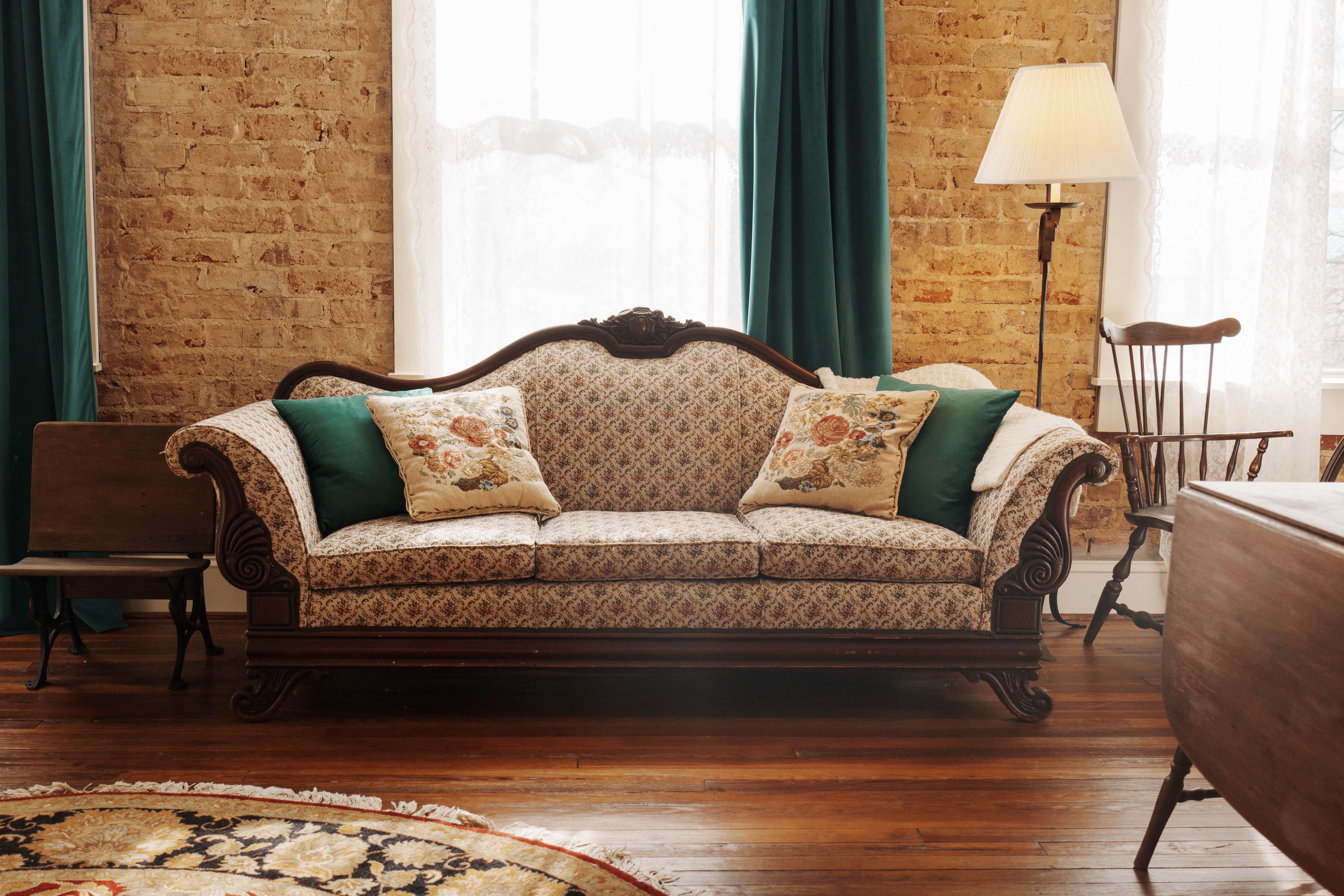 An inviting sitting area with vintage-style sofa with pillows, a floor lamp, and a wooden chair in a room with brick walls and teal curtains.