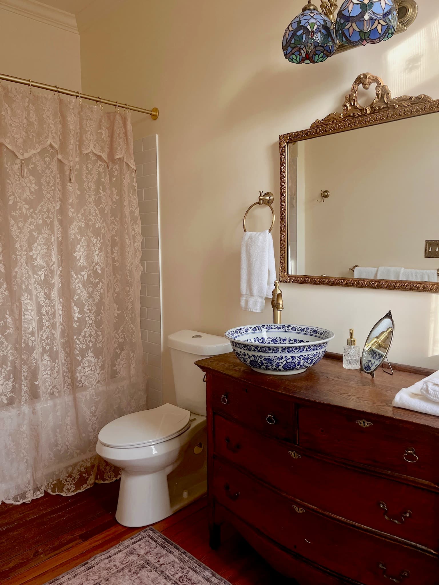 A bathroom with a dark wooden dresser-style vanity, a blue and white patterned bowl sink, a white toilet, and a shower with a lace curtain.