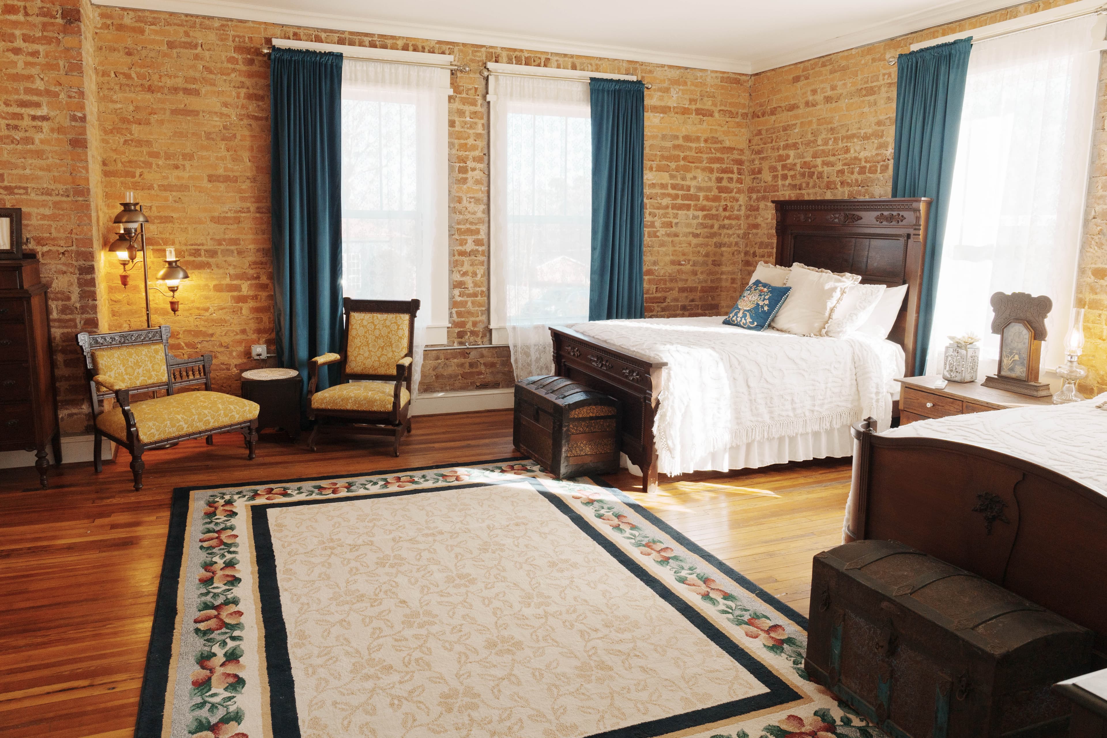A bedroom with two full beds featuring white linens, two chairs, a patterned rug, brick walls, and teal curtains.