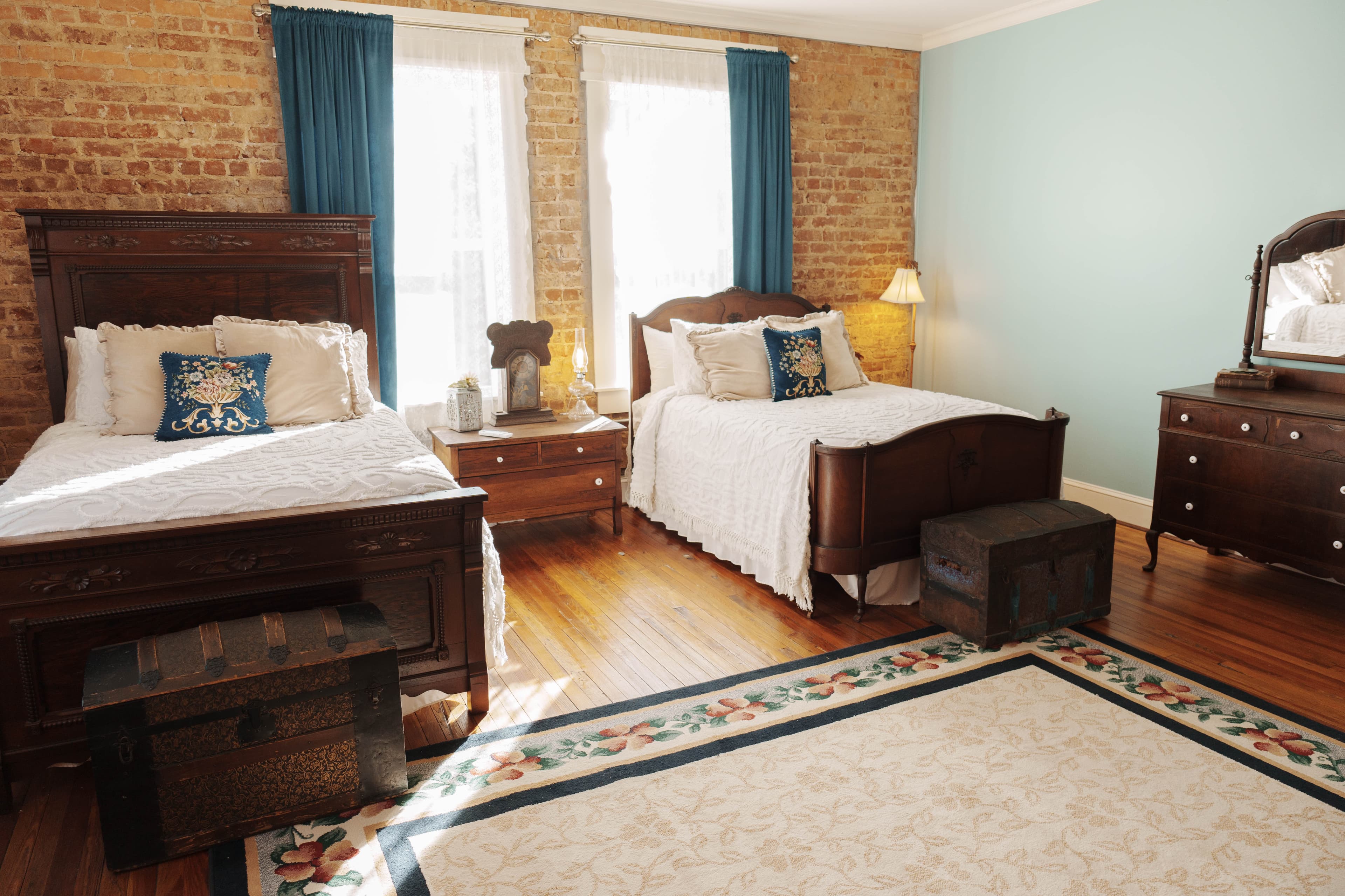 A bedroom with two wooden full beds with white linens, a wooden dresser, a patterned rug, brick walls, and teal curtains.
