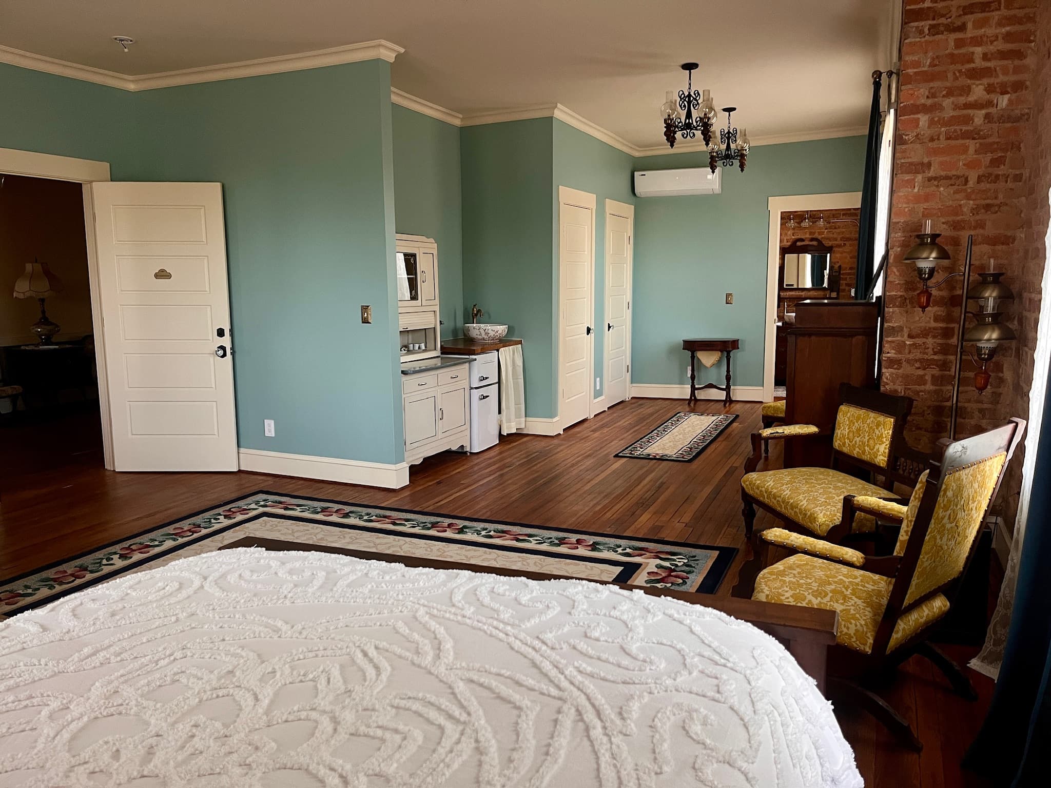 A bedroom with a bed featuring a white chenille spread in the foreground, two upholstered chairs, a patterned rug, blue walls, exposed brick, and a kitchenette.