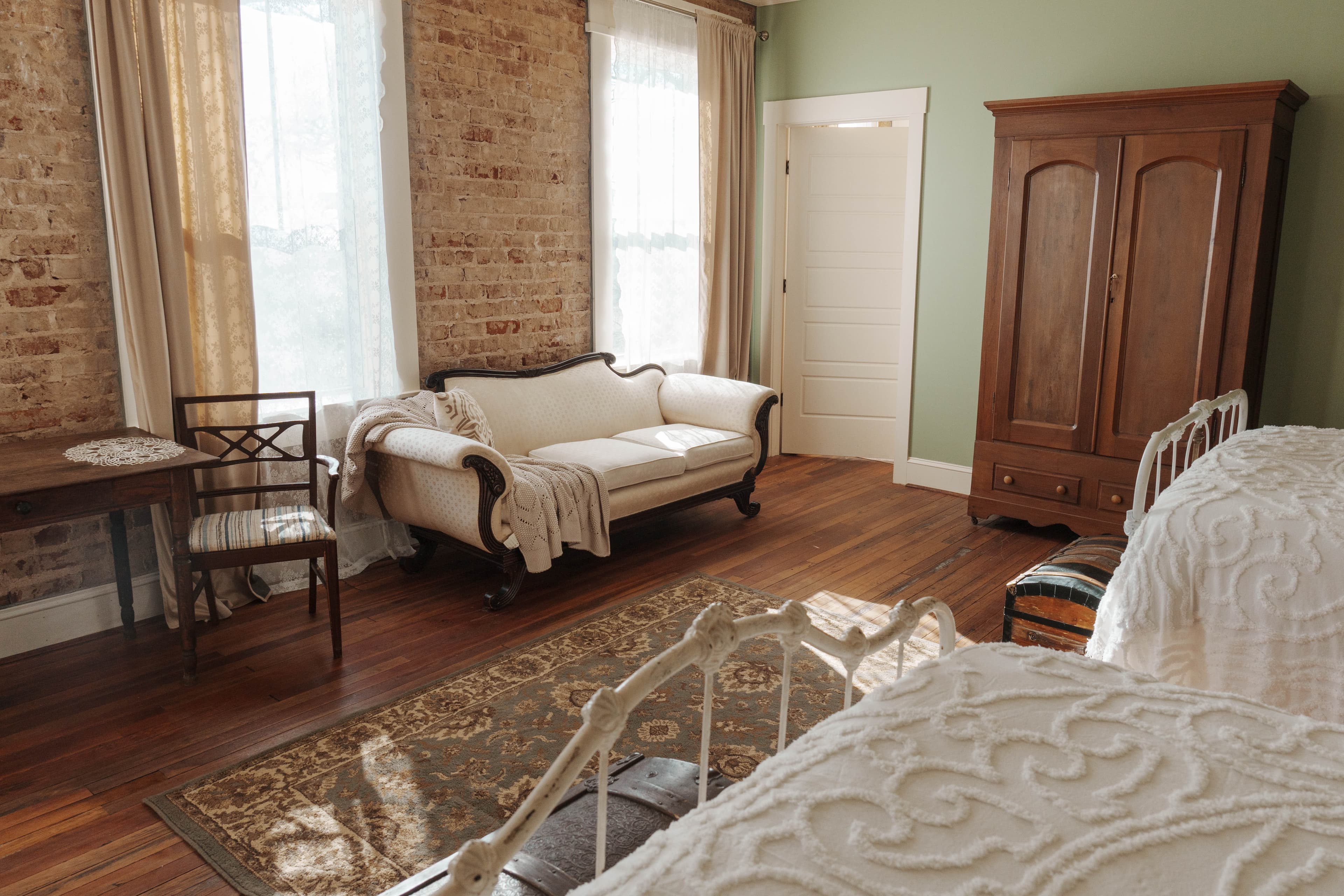 Cozy sitting area opposite beds with vintage-style sofa, wooden table with two chairs, tall sunny windows and exposed brick walls.