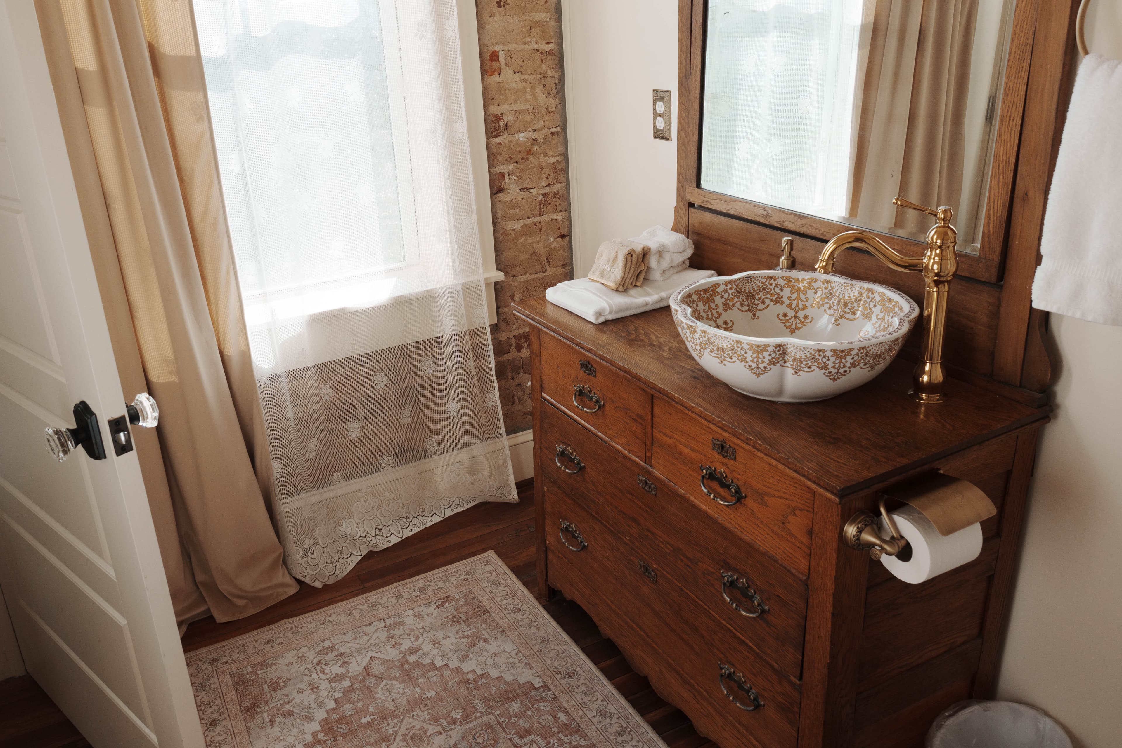 Bathroom with dresser-style vanity with floral patterned bowl sink, wooden framed mirror, and exposed brick wall.