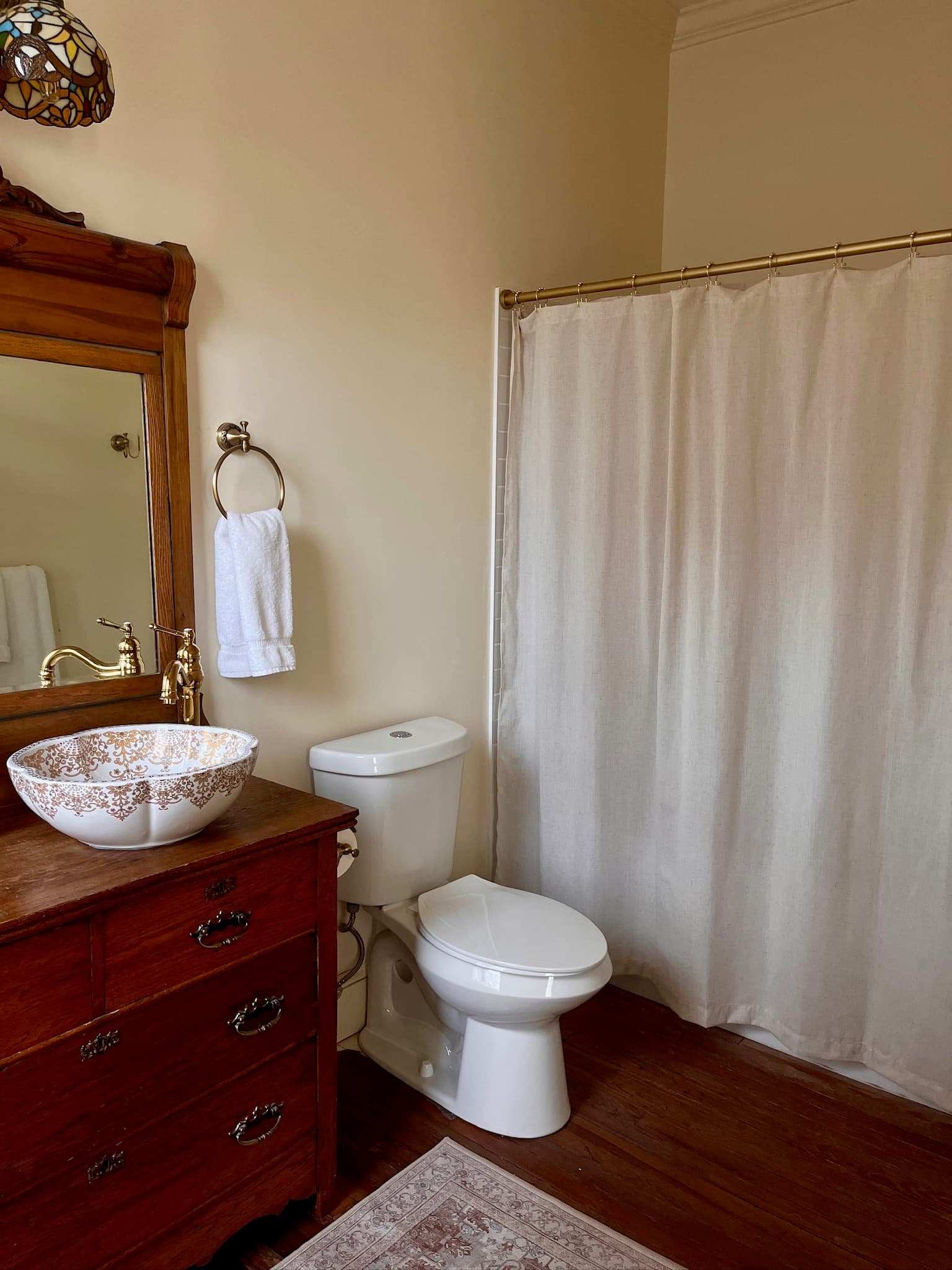 Bathroom with dresser-style vanity with floral patterned bowl sink, wooden framed mirror, toilet, and shower.
