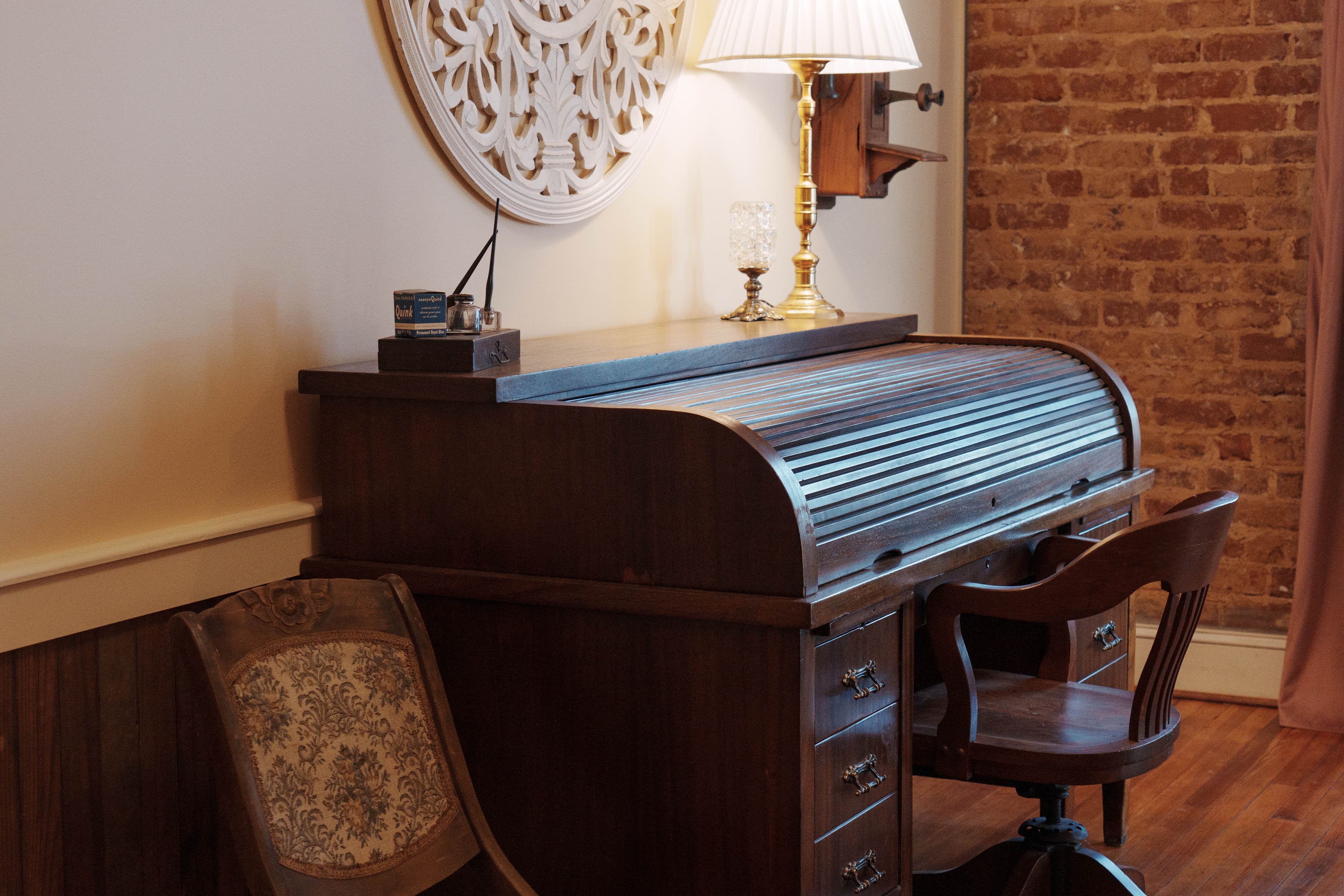 A vintage wooden roll-top desk with a matching chair, a lamp, and decorative elements against a brick wall.