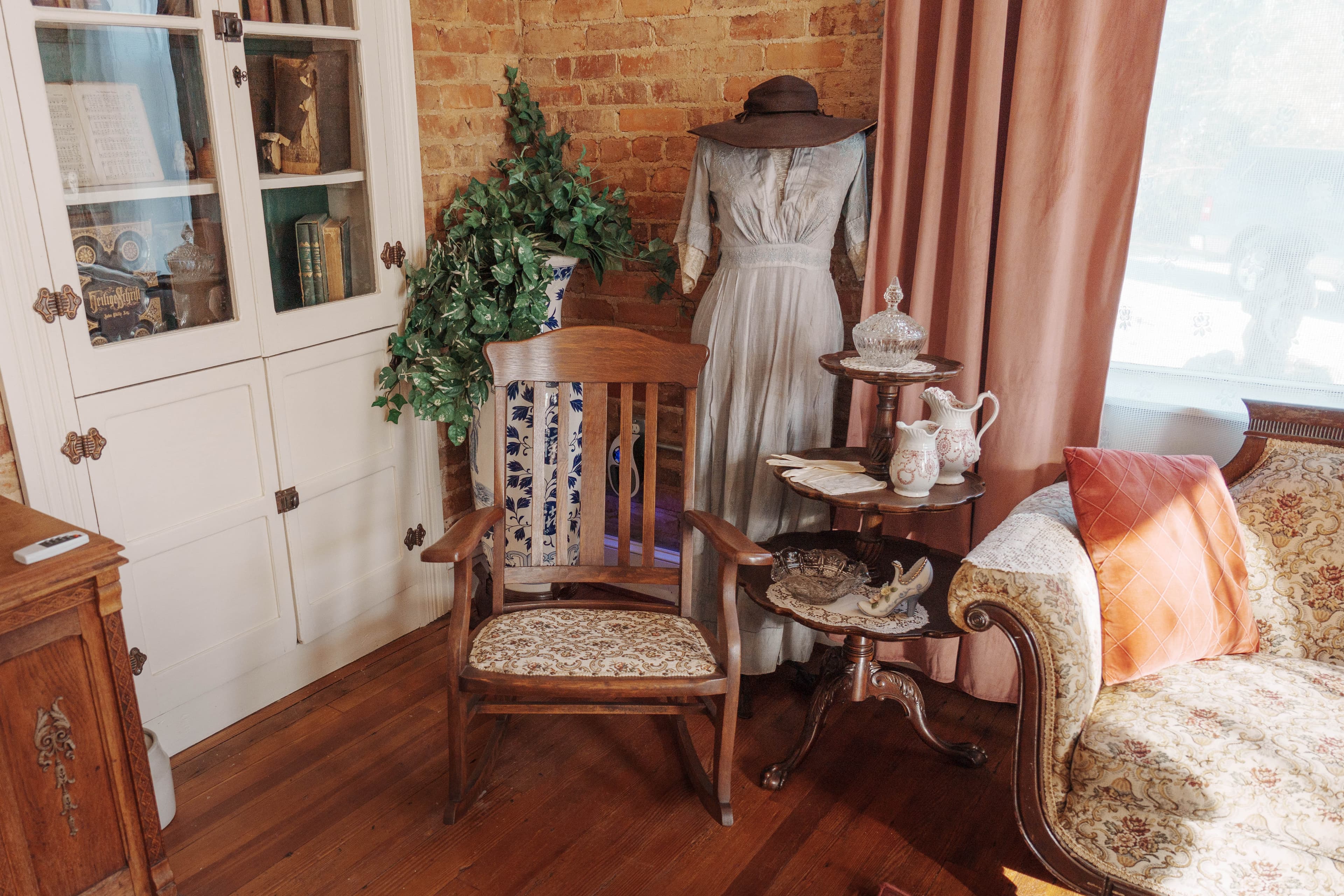 A cozy vintage living room featuring a wooden chair, a decorative table with fine china, and a mannequin dressed in an elegant gown.