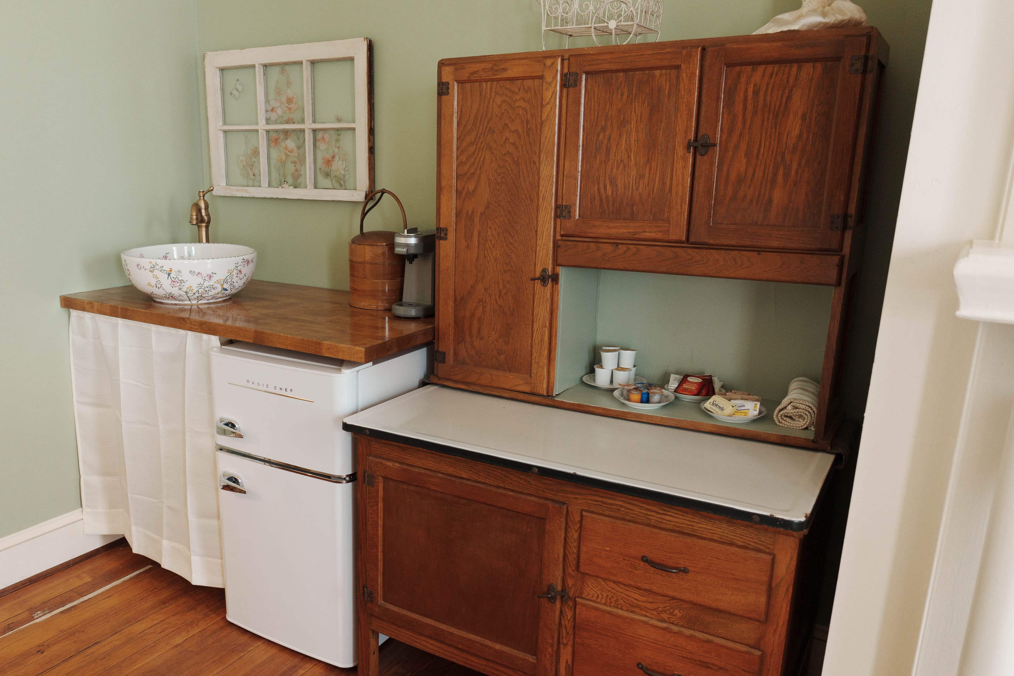 A rustic kitchen corner featuring a wooden countertop, vintage storage cabinets, a white sink, and a small refrigerator.