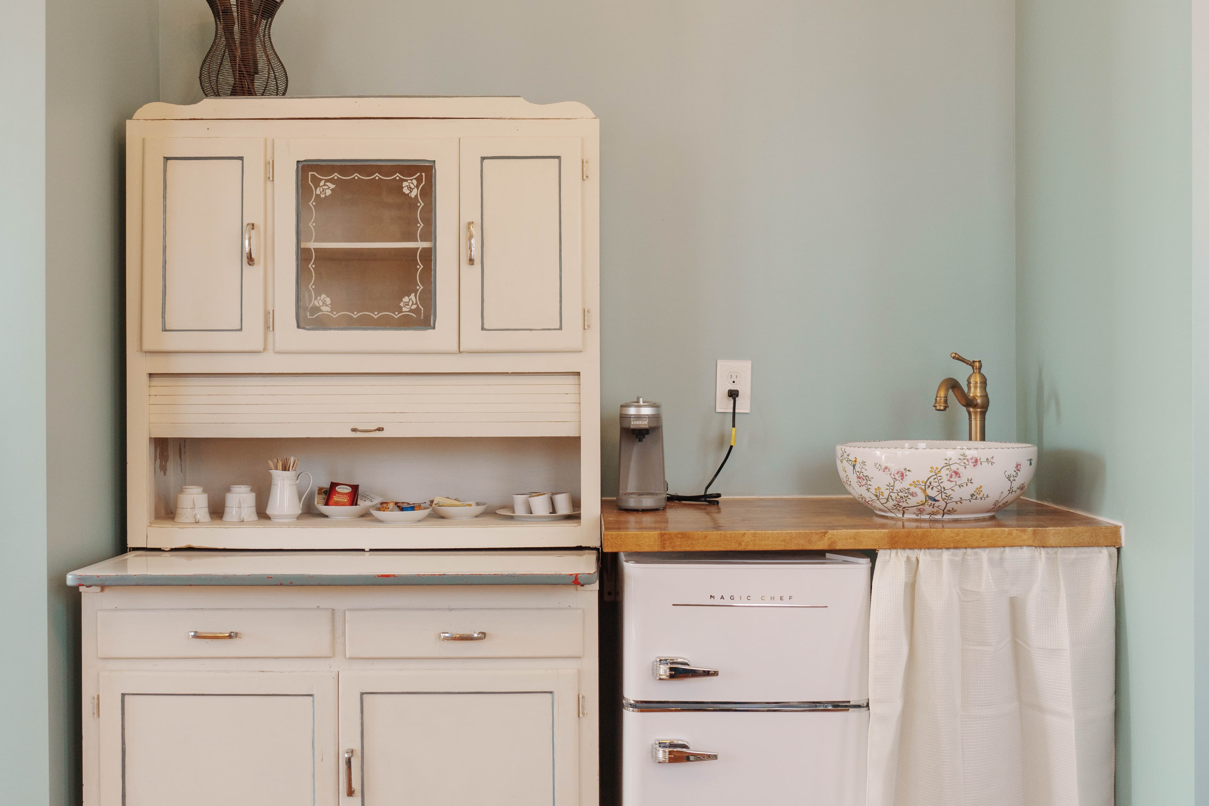 A vintage-style kitchen corner featuring a white hutch, a small refrigerator, and a countertop sink with decorative bowl.