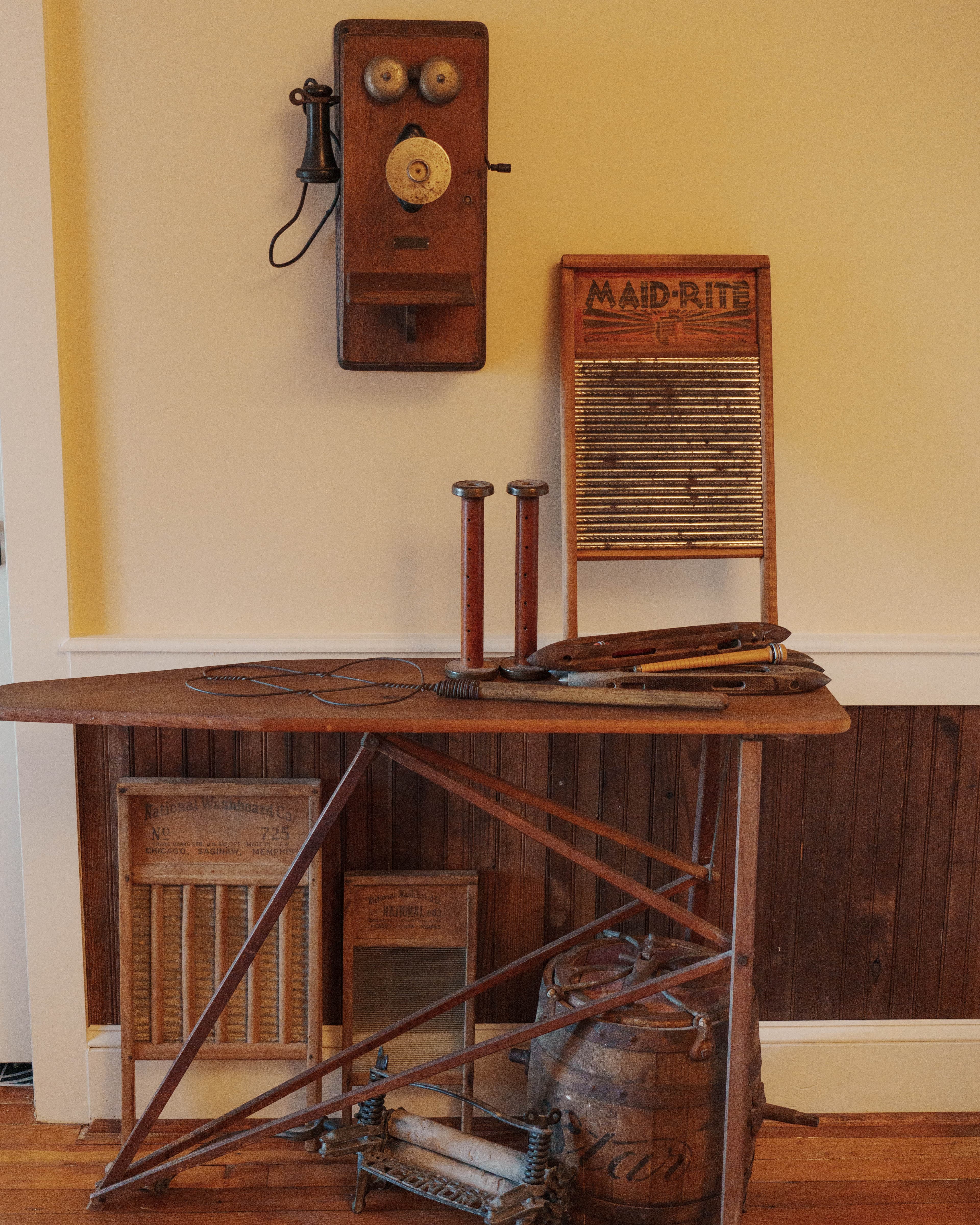 A vintage wooden table displays an antique telephone, washboard, and various old tools against a neutral wall.