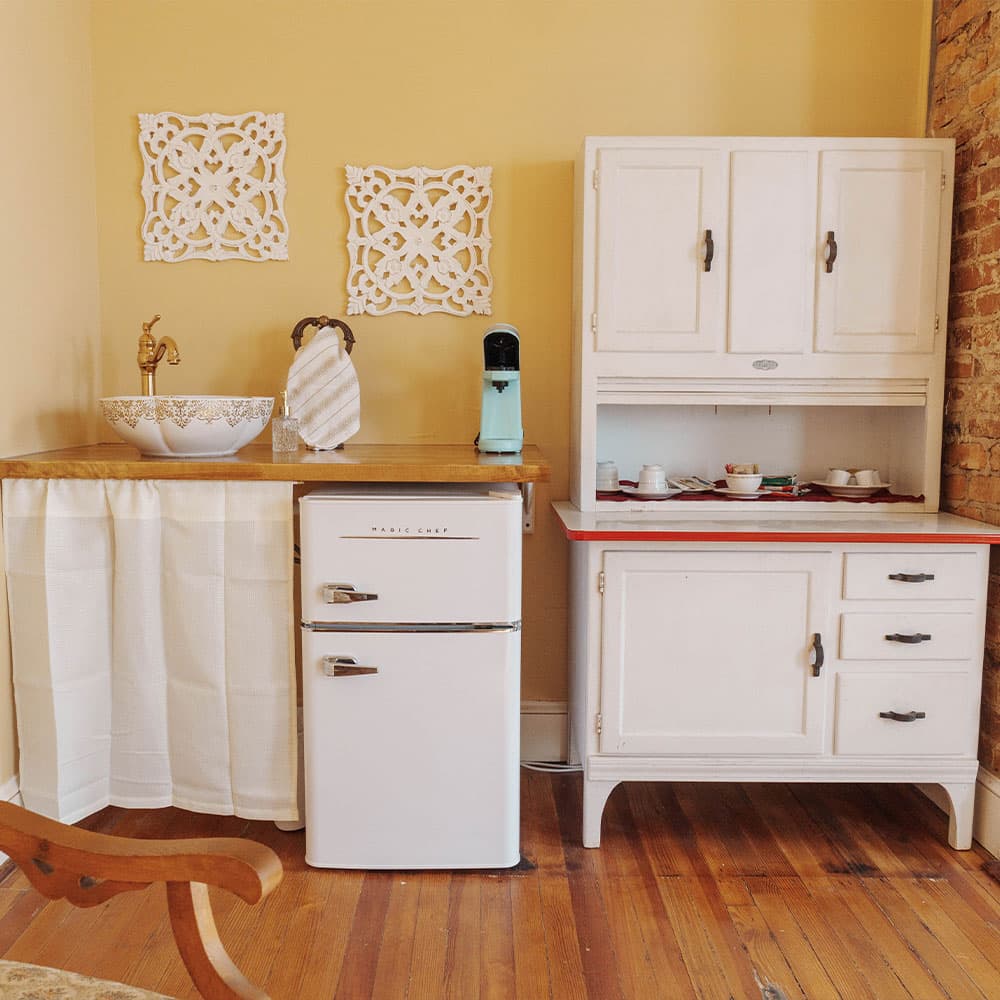 A quaint kitchen area featuring a white refrigerator, a wooden countertop with a sink, and vintage cabinetry against a yellow wall.