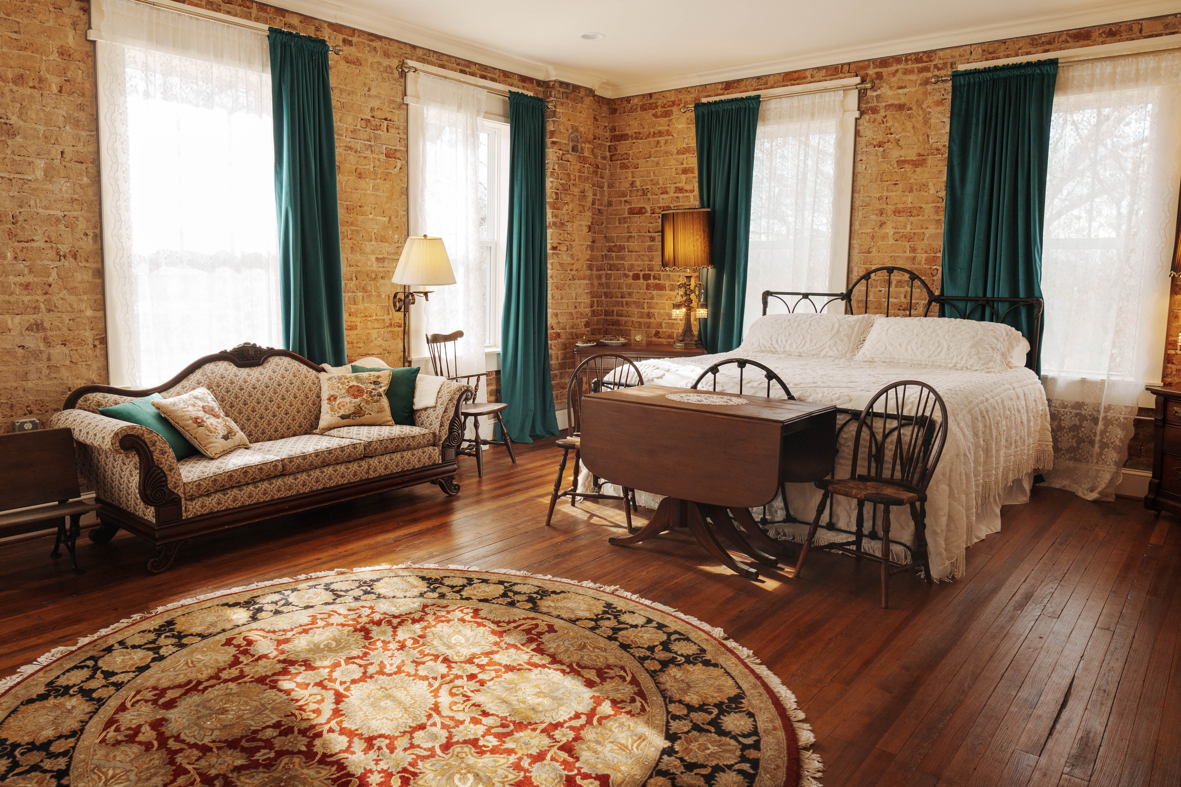 A bedroom with a king-size bed, a beige sofa with decorative pillows, a round patterned rug, and multiple windows with teal curtains.