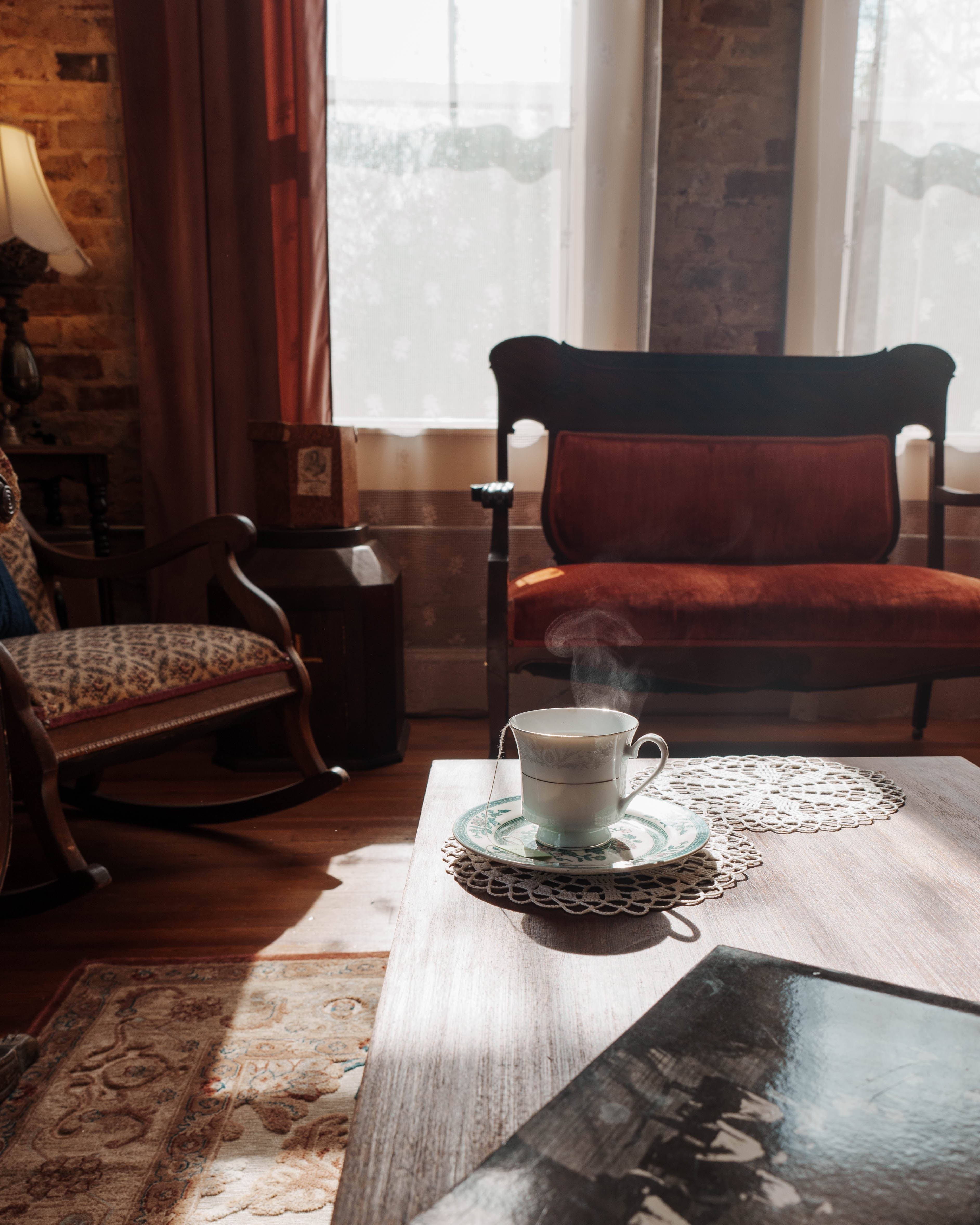 A steaming cup of tea sits on a lace doily atop a wooden table, with a cozy room in the background.