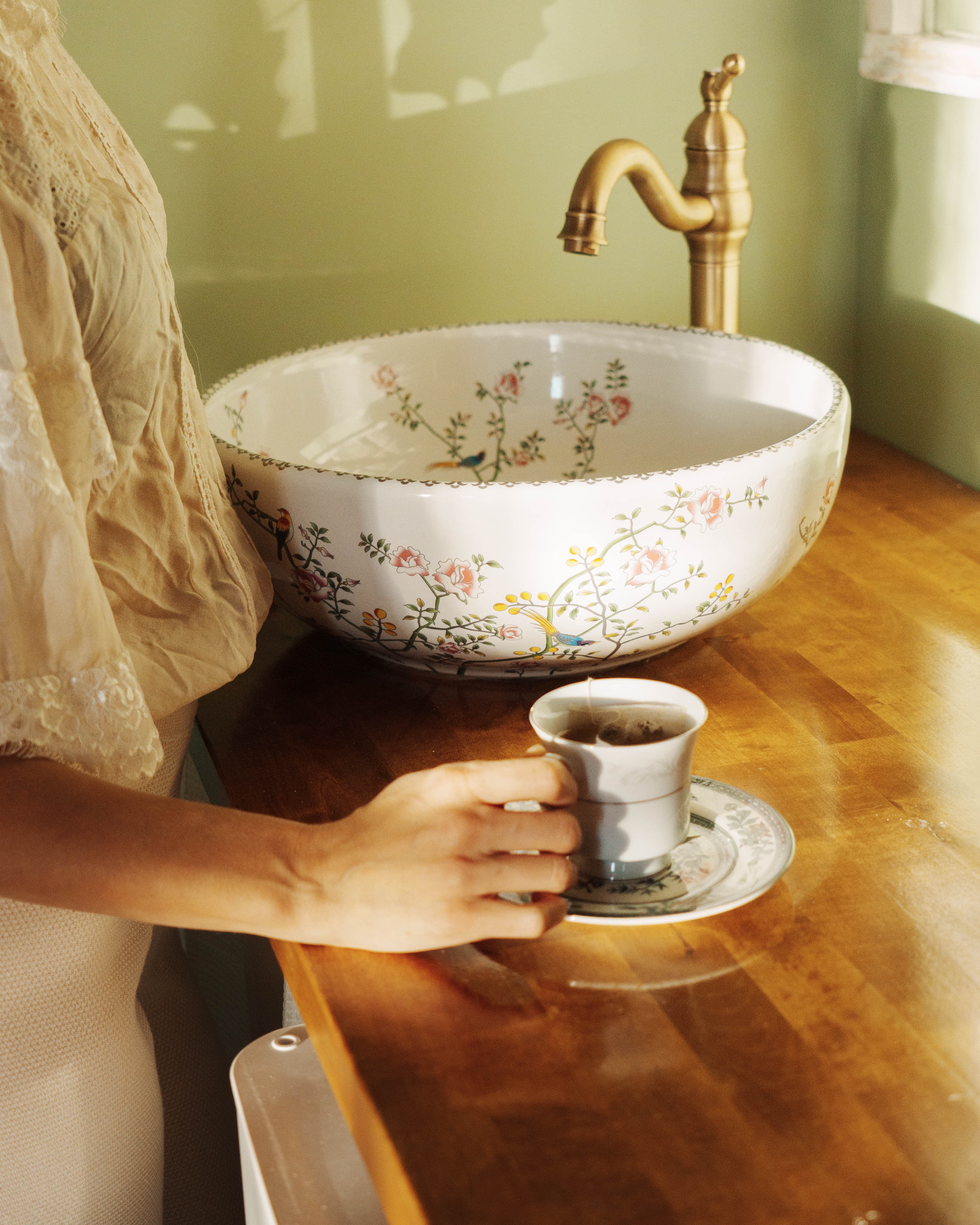 A hand holds a small cup of tea next to a floral-patterned bowl and a brass faucet on a wooden countertop.