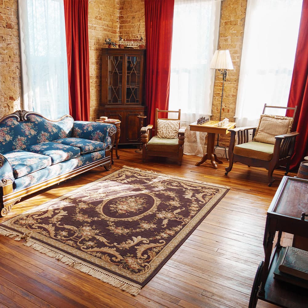 A cozy living room featuring a floral couch, two wooden chairs, a coffee table, and a decorative rug on hardwood floors, illuminated by natural light through large windows.