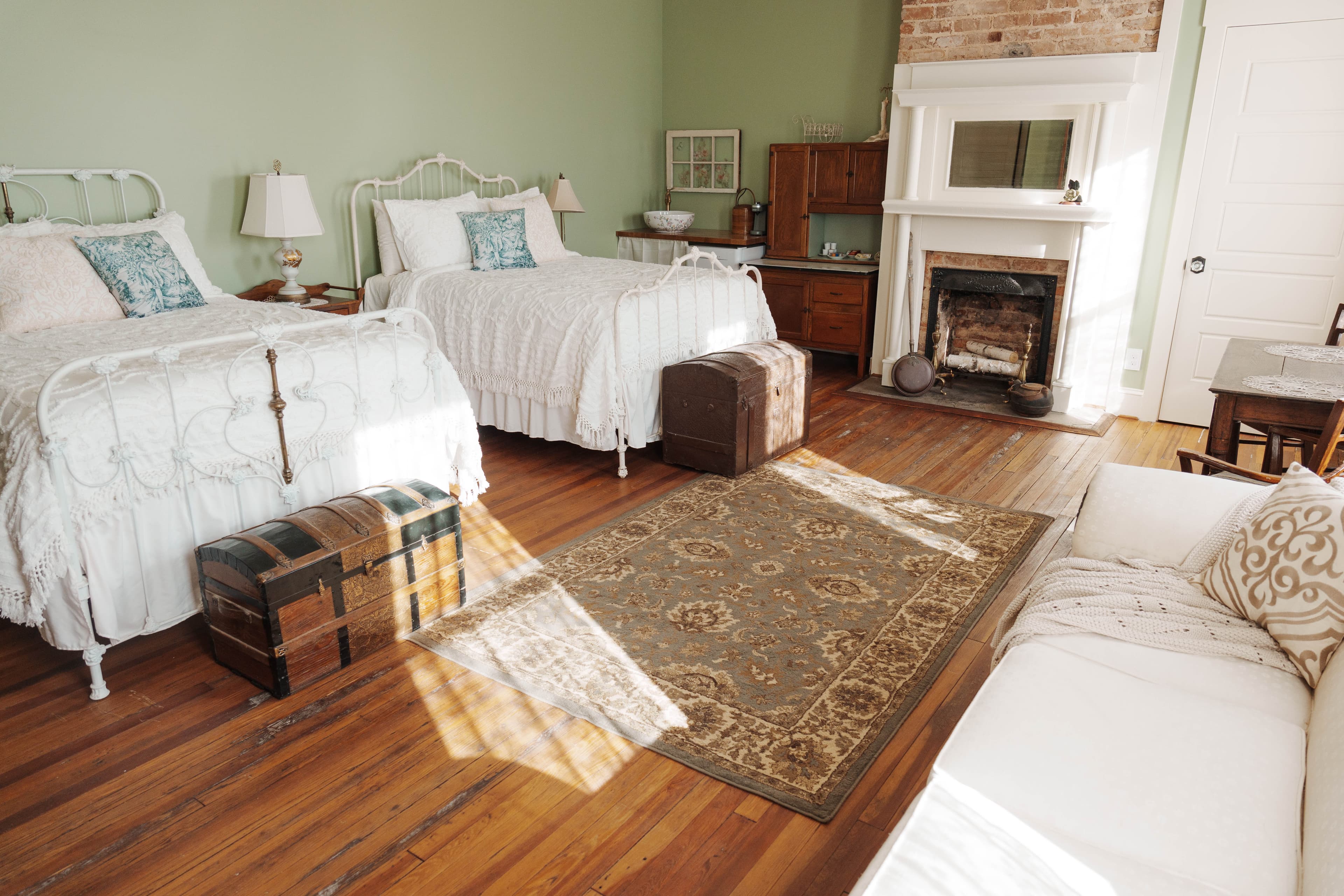 A cozy bedroom with two white metal beds, vintage trunks, a fireplace, and a woven rug on wooden flooring.