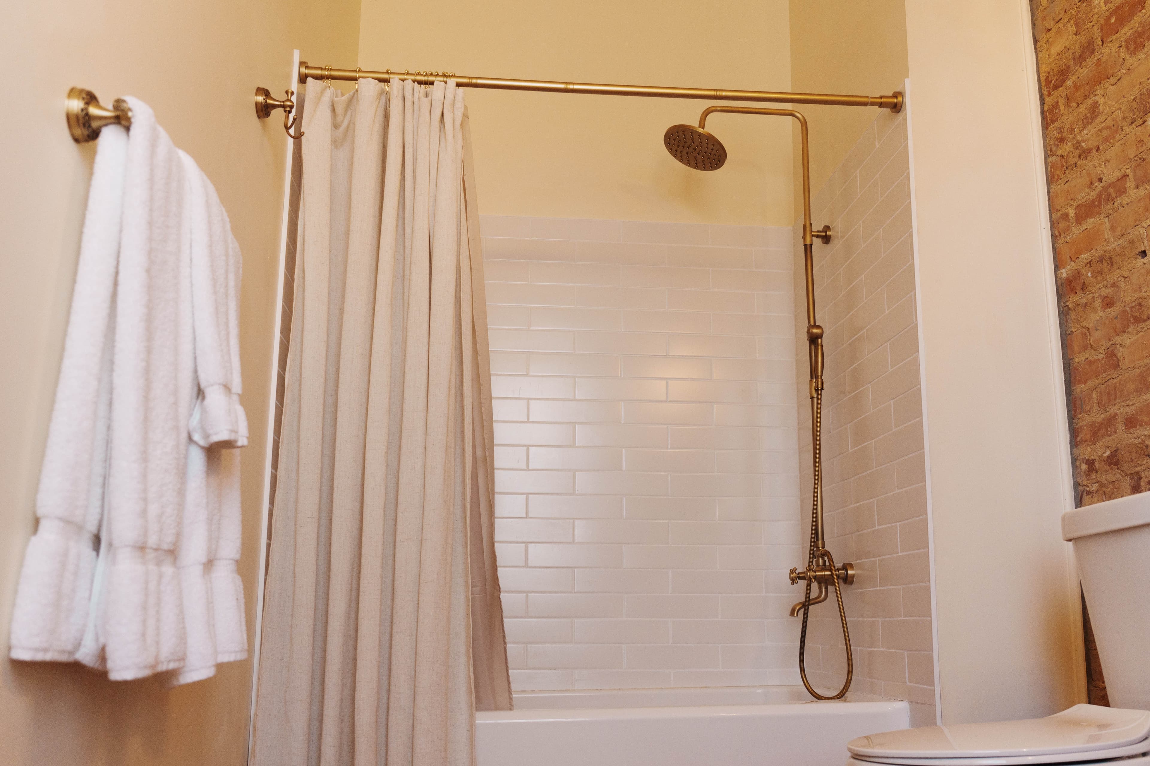Bathroom with tiled shower/tub combination, rain showerhead, and toilet with exposed brick walls. Bright white plush towels hang at the ready on a rack on the wall.