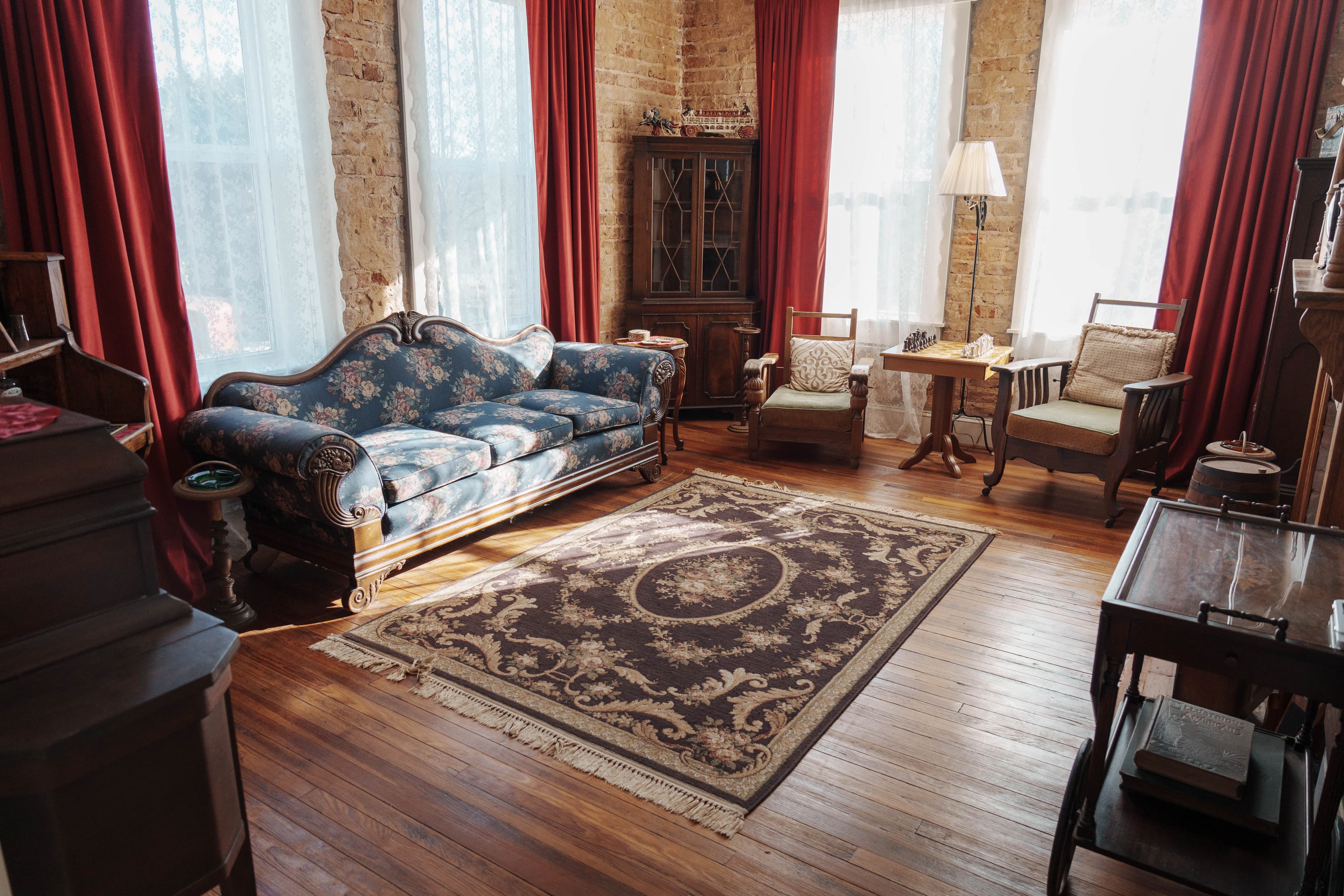 Cozy living room featuring a floral couch, wooden furniture, and large windows with red curtains.