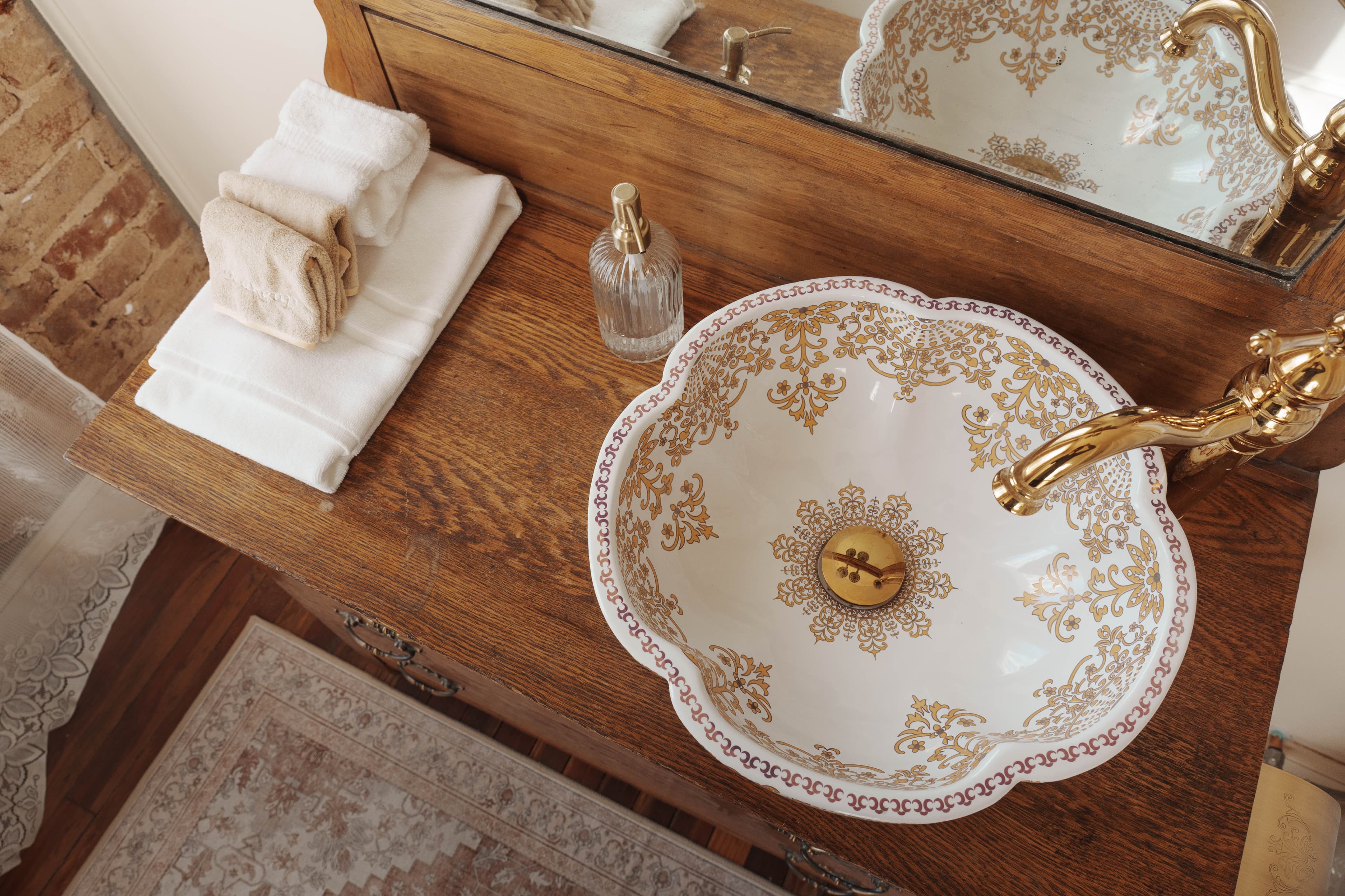 A decorative sink with gold floral patterns sits atop a wooden vanity, accompanied by neatly stacked towels and a glass soap dispenser.