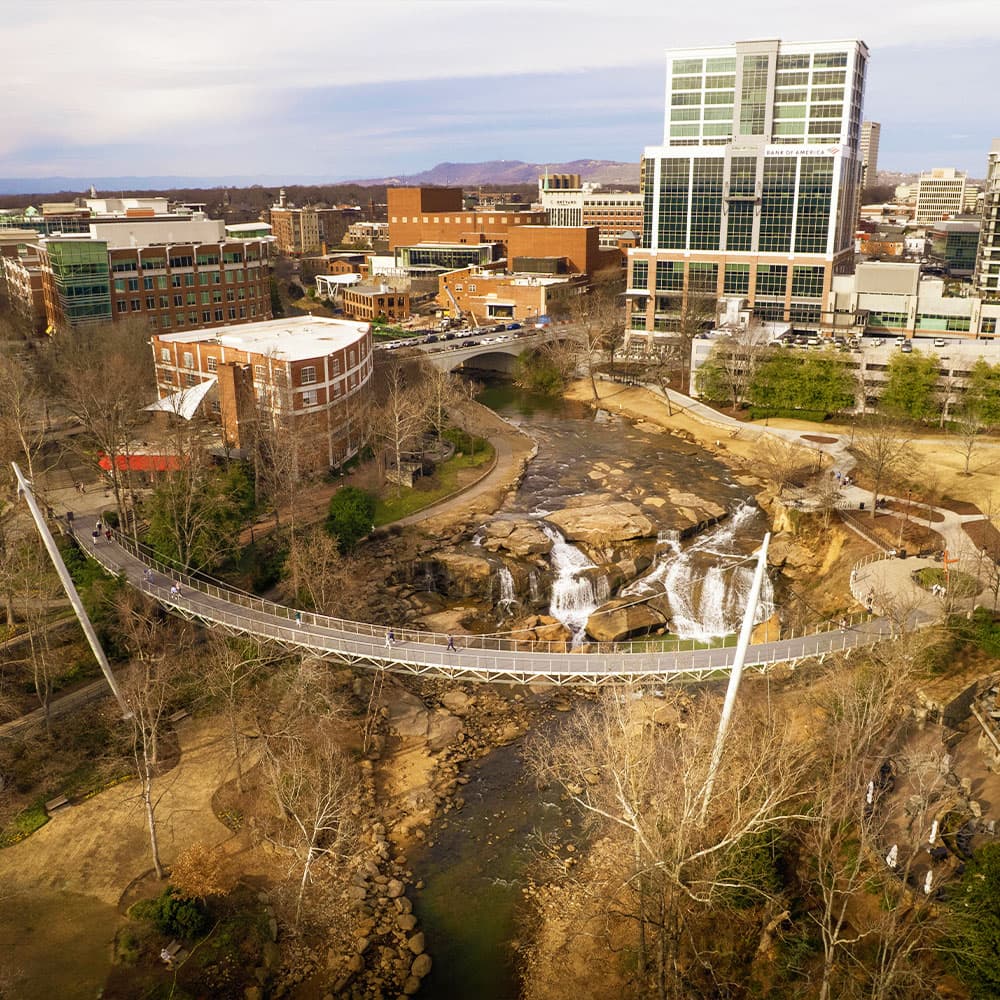 Aerial view of a city park featuring a bridge over a stream with a waterfall, surrounded by buildings.
