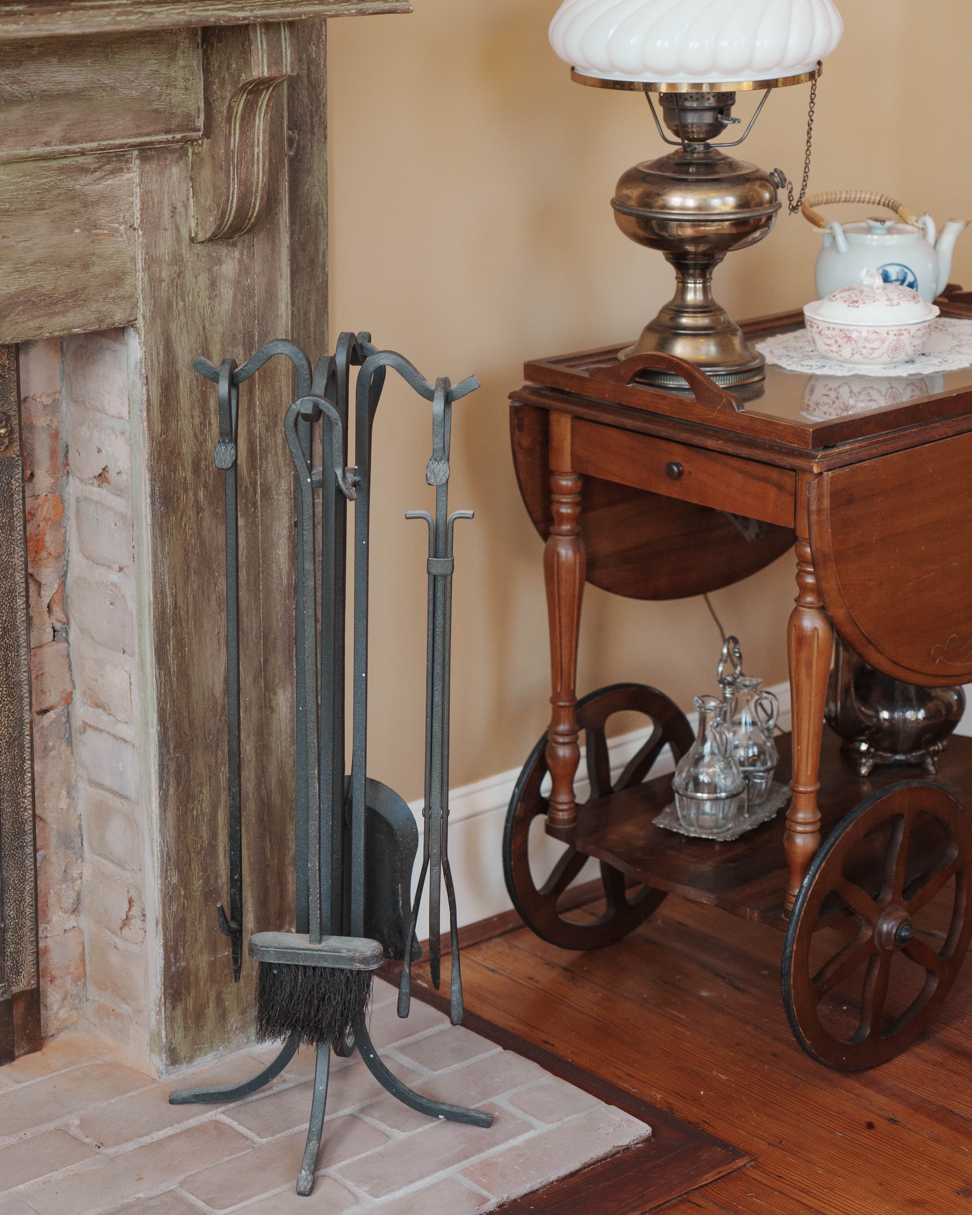 A metal fireplace tool set stands next to a wooden side table with a lamp and decorative items.