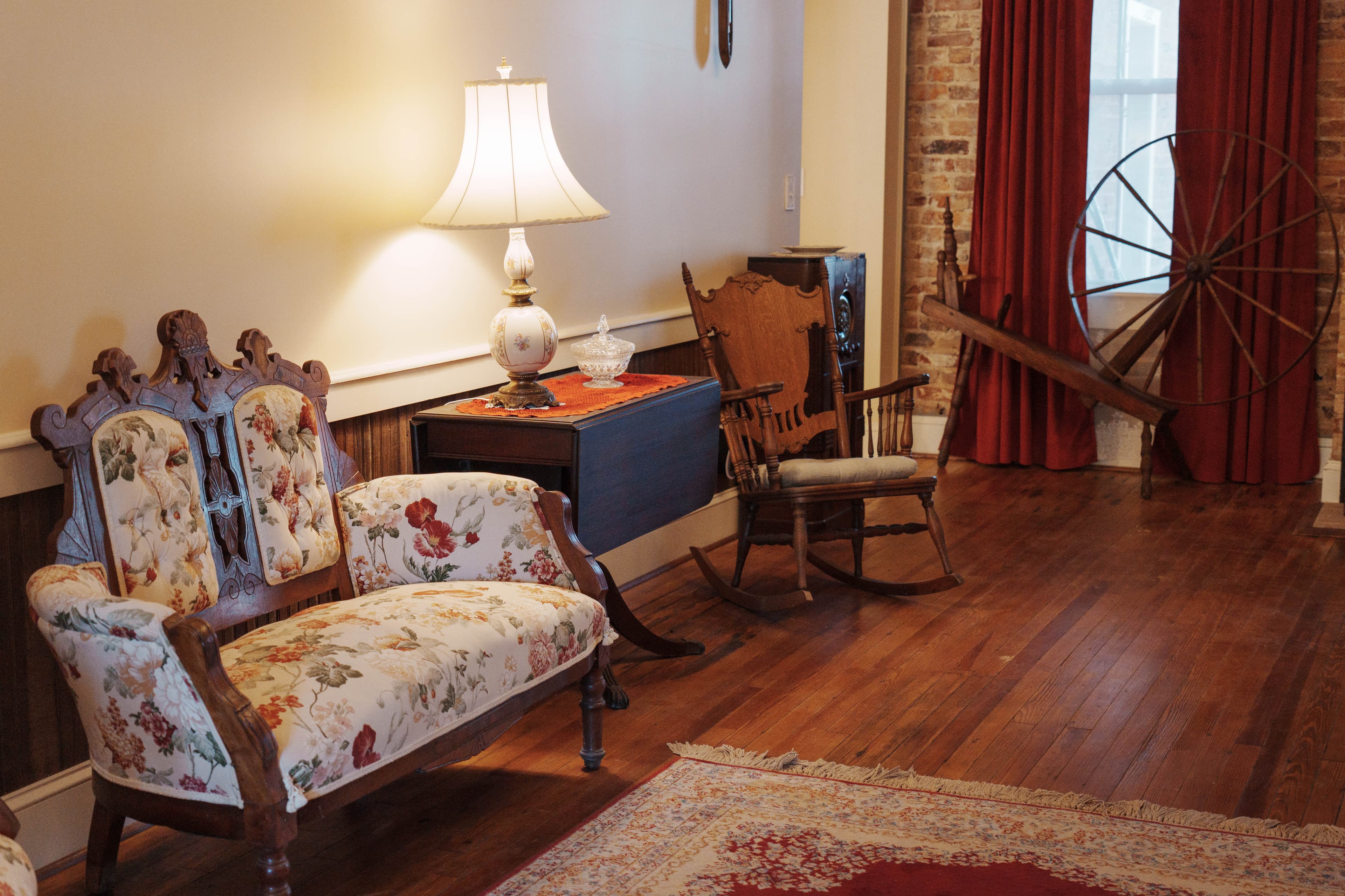 A cozy living room featuring a floral-patterned sofa, a rocking chair, a lamp, and a decorative spinning wheel.