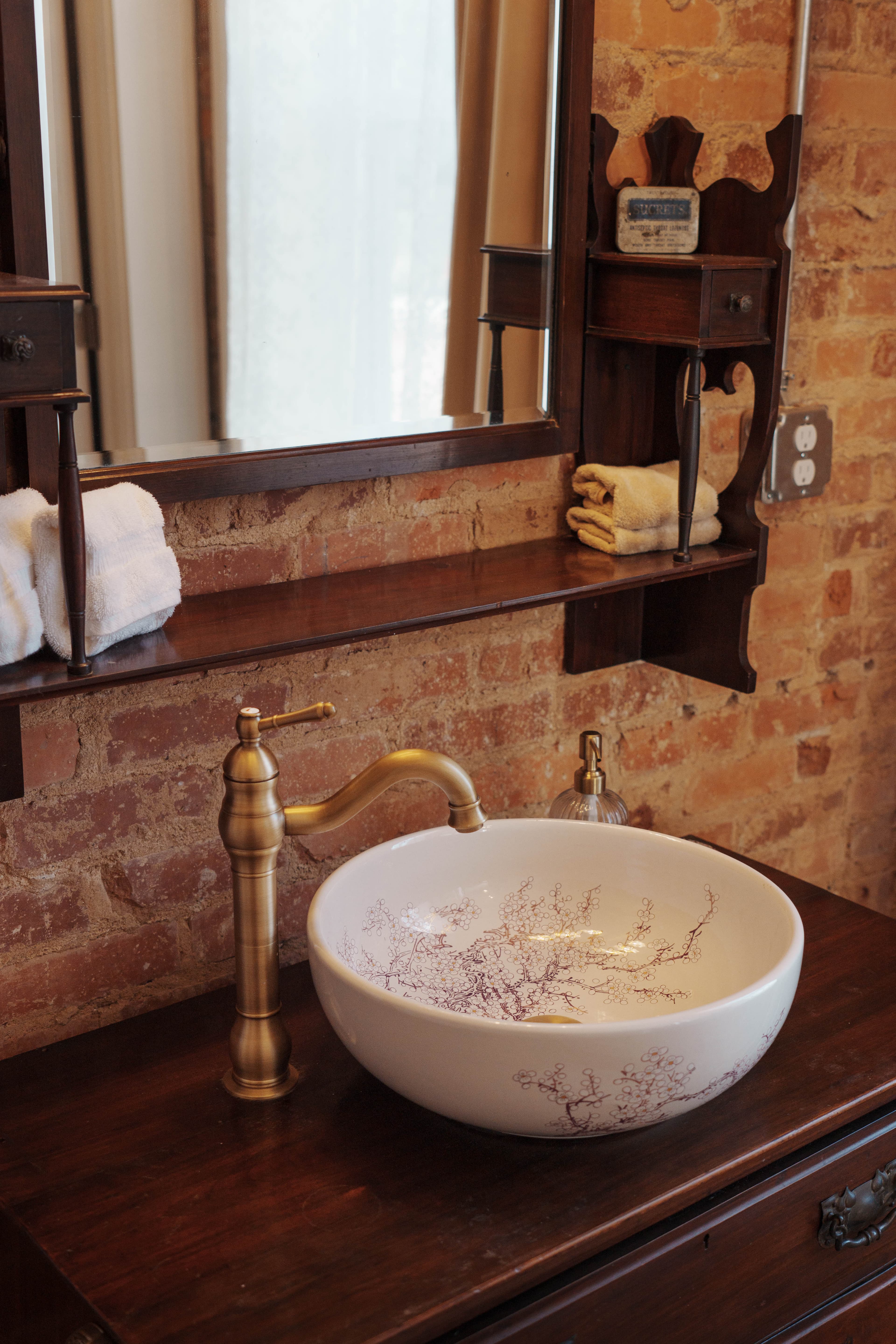 A stylish bathroom sink with a decorative bowl, gold faucet, and wooden shelf against a brick wall.