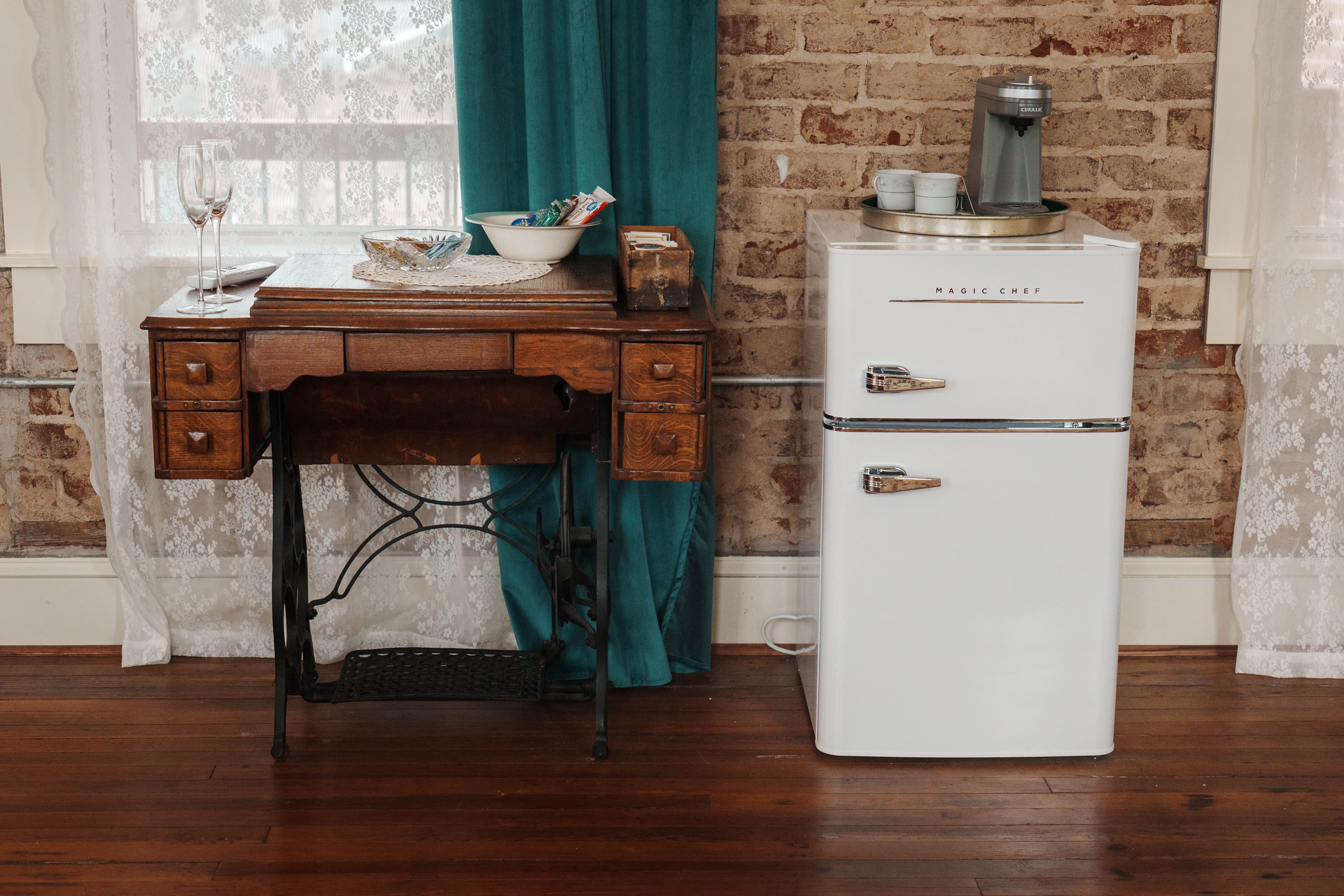 A vintage wooden desk and a retro white refrigerator are positioned against a brick wall, with lace curtains and turquoise accents in the background.
